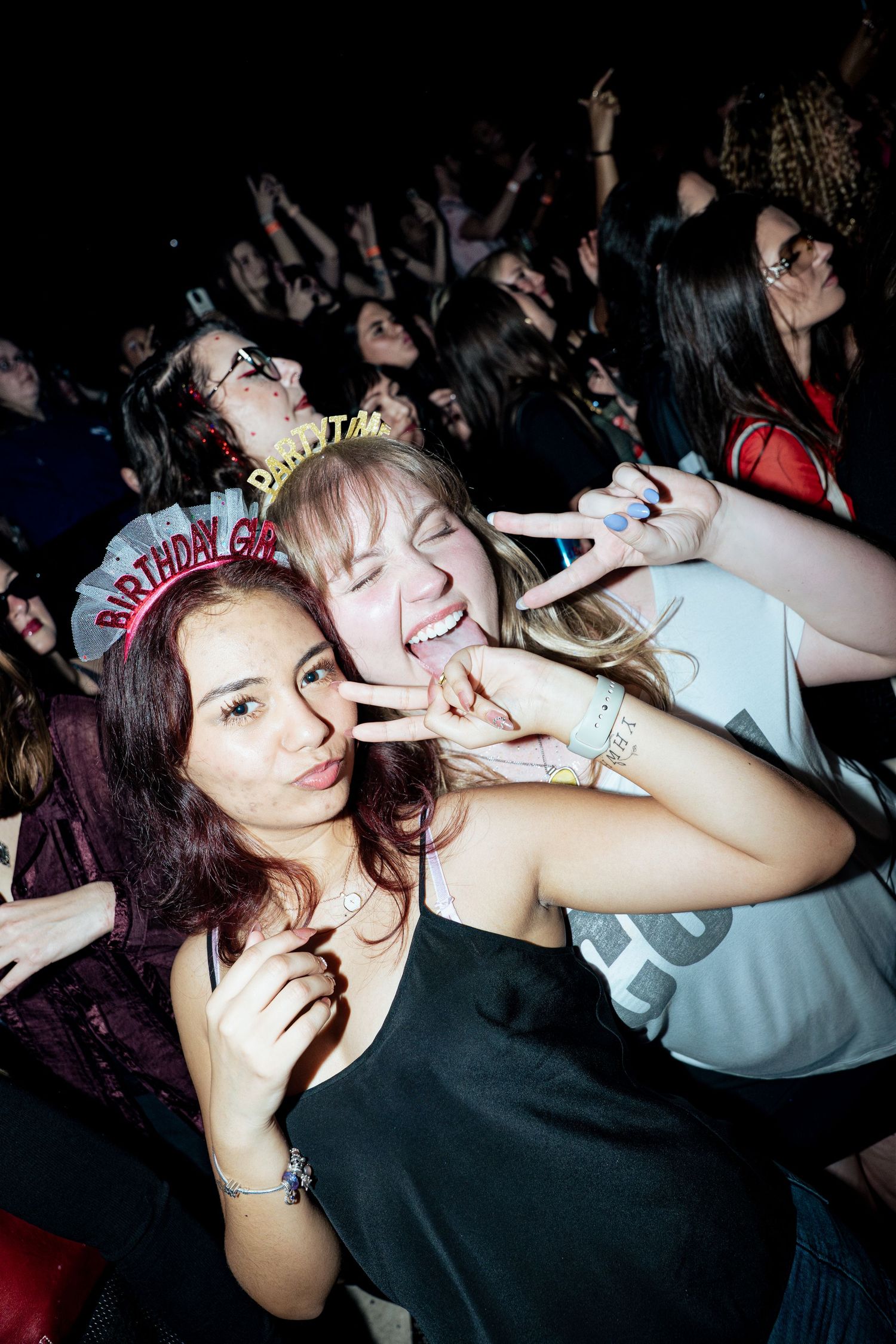 Two young women celebrating a birthday at a concert, making peace signs, one wearing a Birthday Girl tiara.