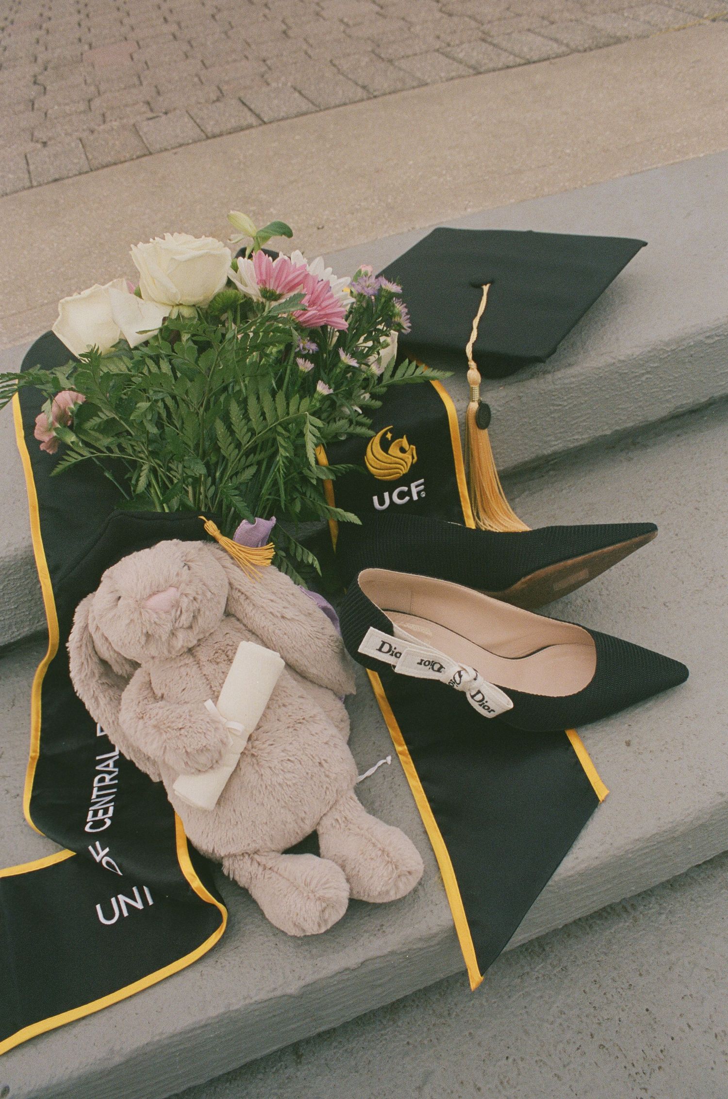 UCF graduation cap, green sash, bouquet of flowers, stuffed bunny, and black heels arranged on concrete steps.