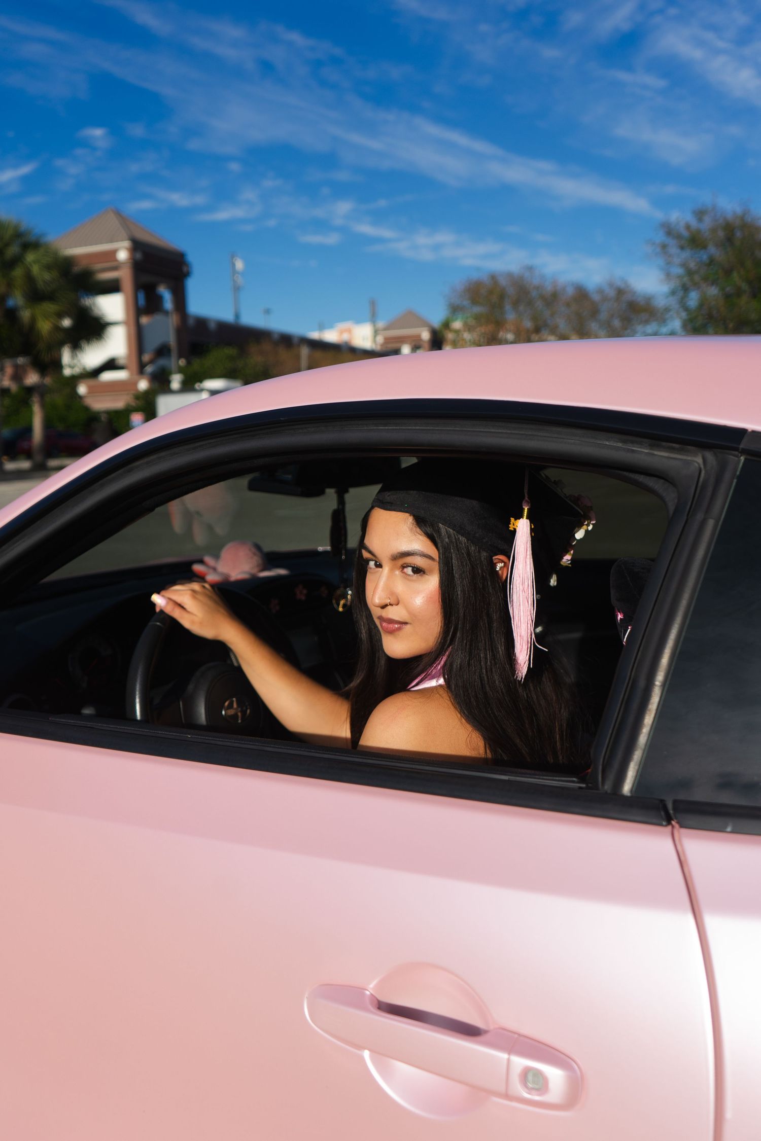 Female graduate wearing cap and tassel poses in a pink car on a bright sunny day.