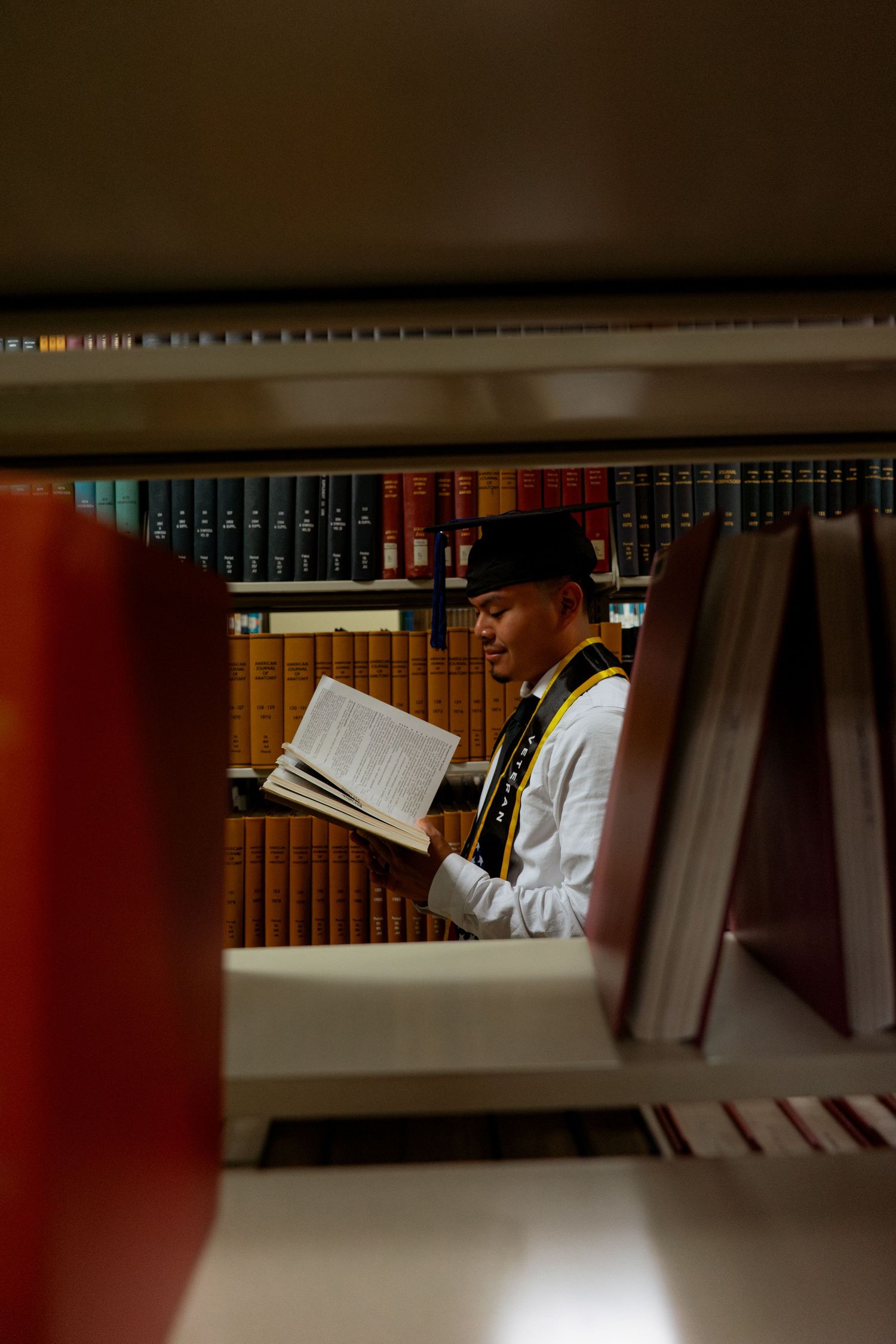 Man in traditional hat reads book in library aisle, surrounded by shelves of antique books, viewed through gap in shelves.