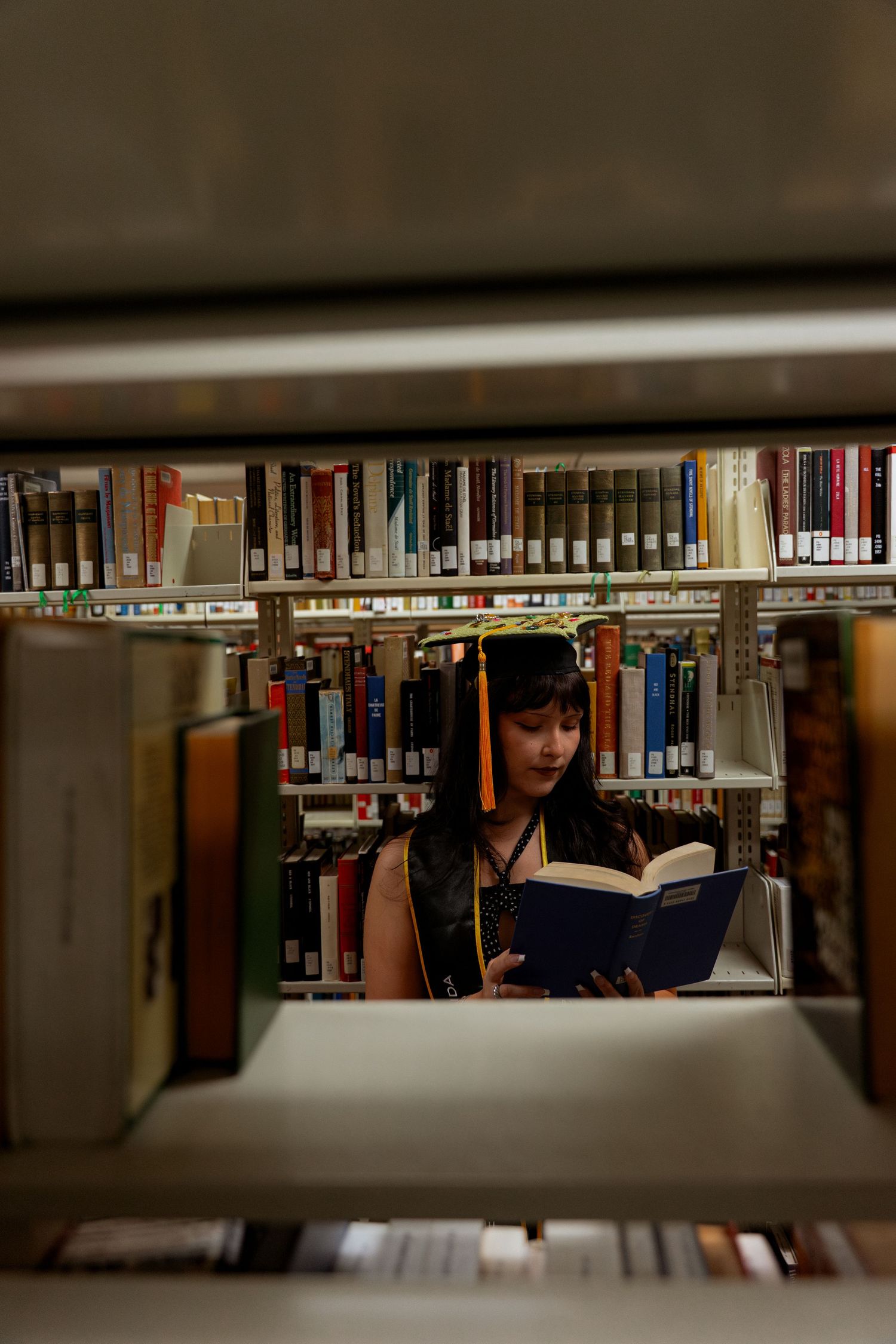 A young woman reads a book in a library, framed by shelves of colorful books in the foreground and background.