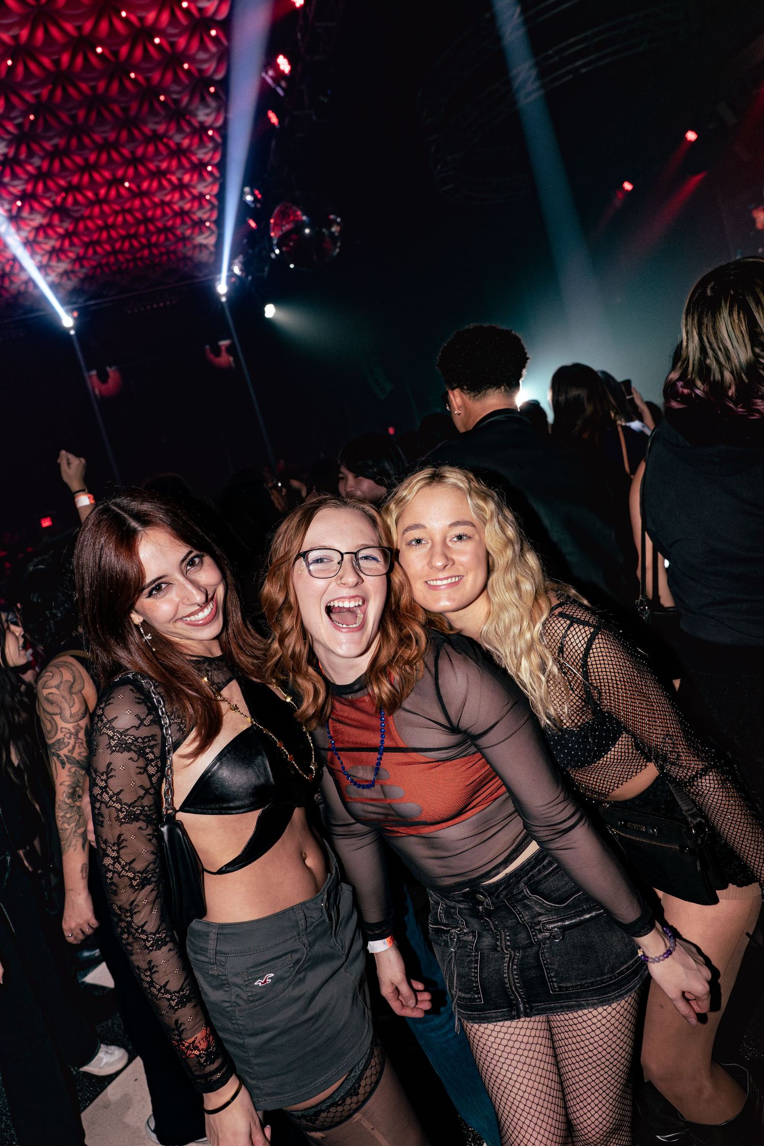 Three smiling women posing together in stylish outfits under red lights at a nightclub event.