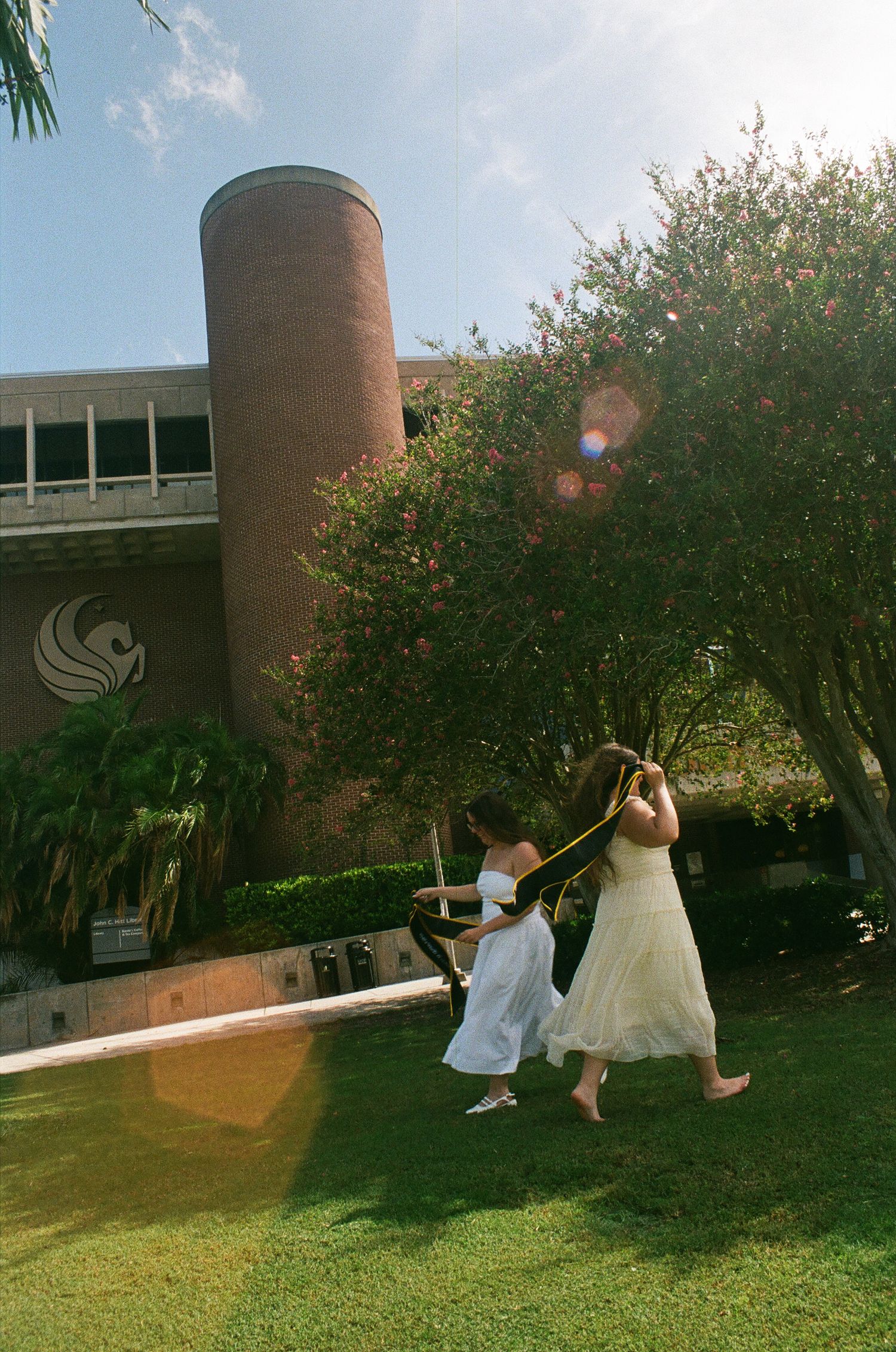 Two women in white dresses dance on green lawn near a brick building with a cylindrical tower on a sunny day.