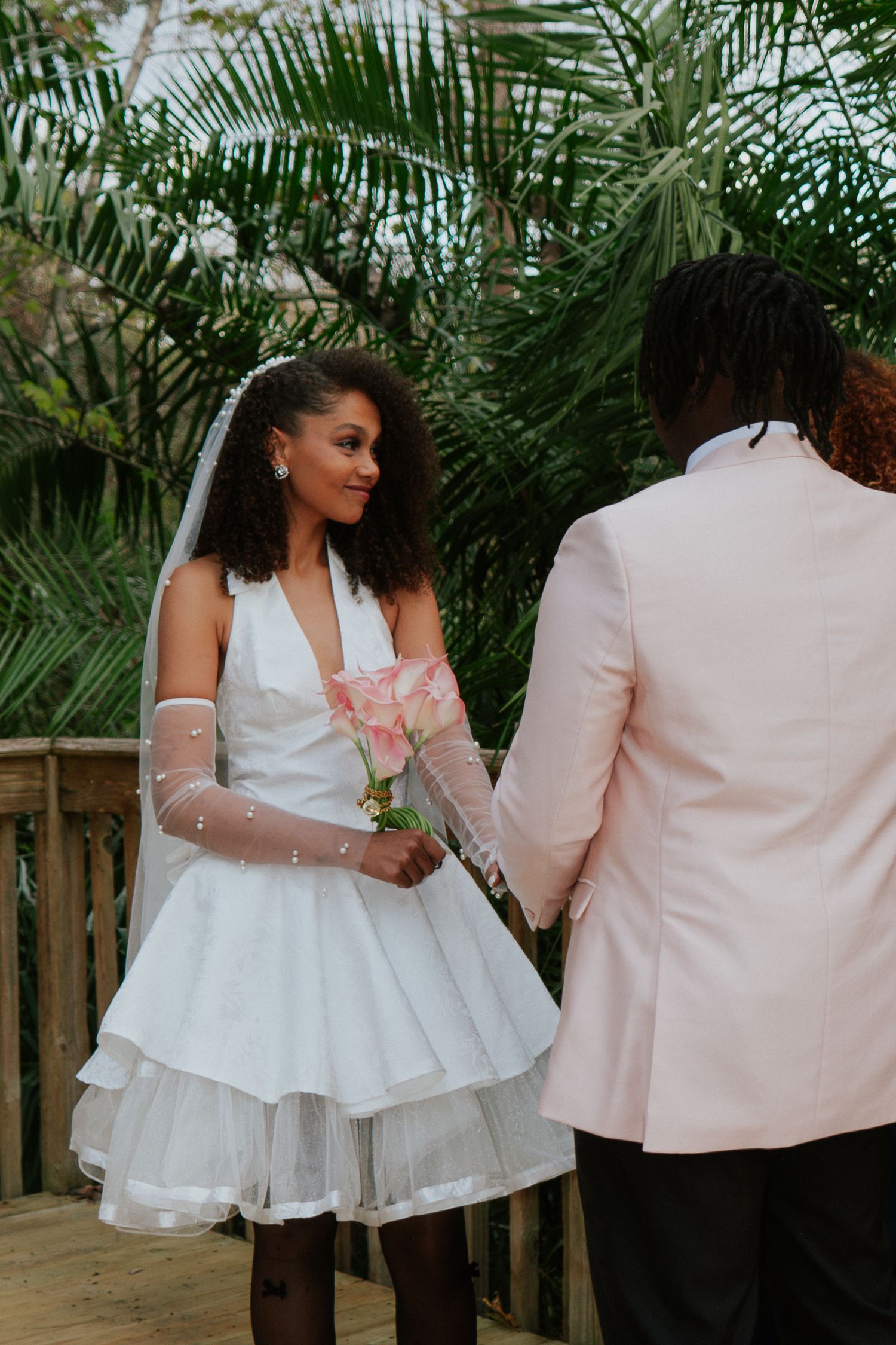 Bride in short white halter dress holds pink roses while groom in pink blazer holds her hand outdoors.