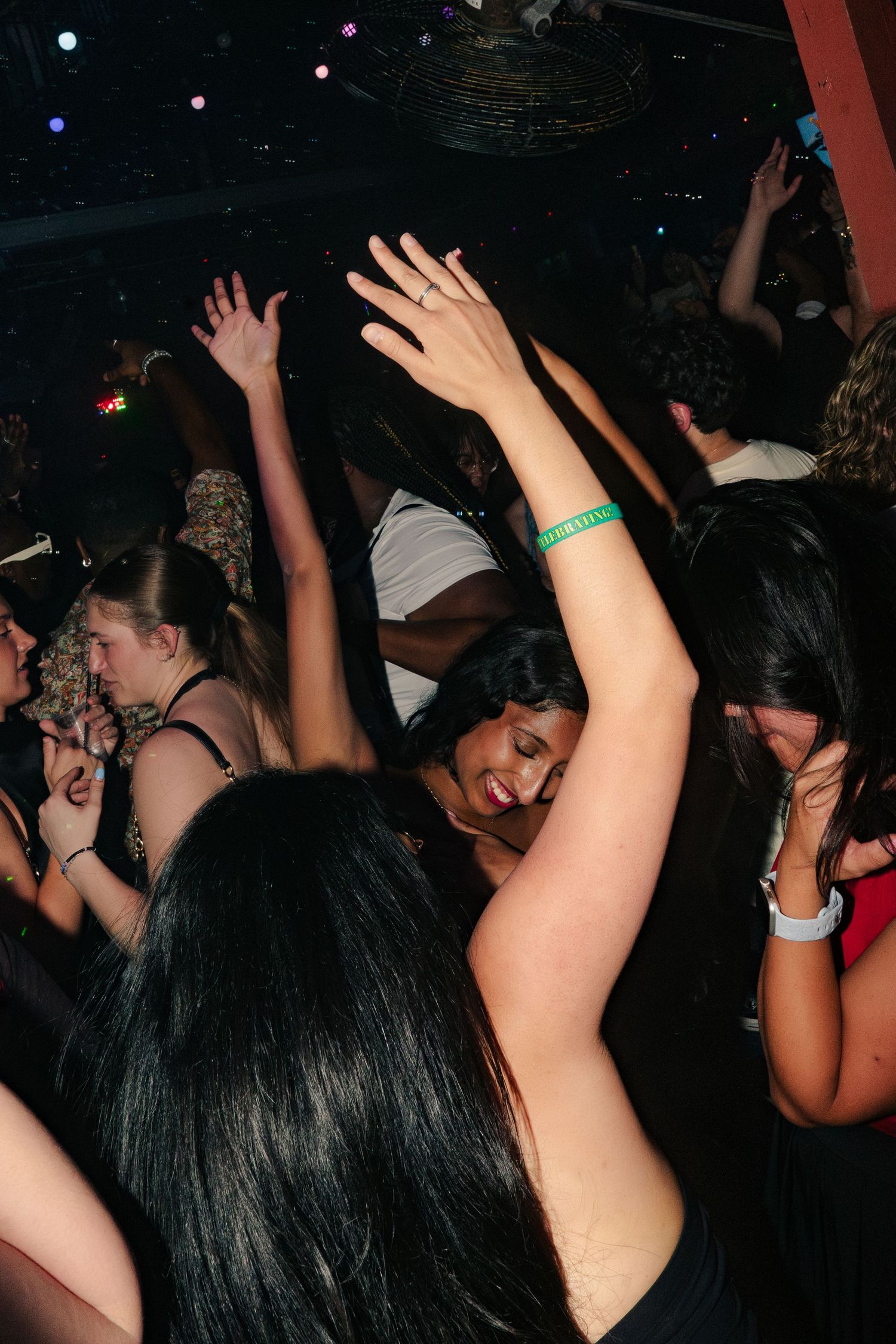 Young women dancing with hands raised at a crowded nightclub party under dim lights.