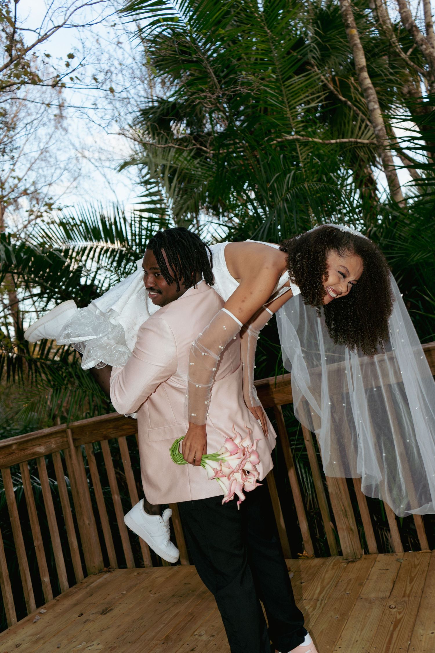 Groom in pink suit carries laughing bride in white dress on his back on a wooden deck.