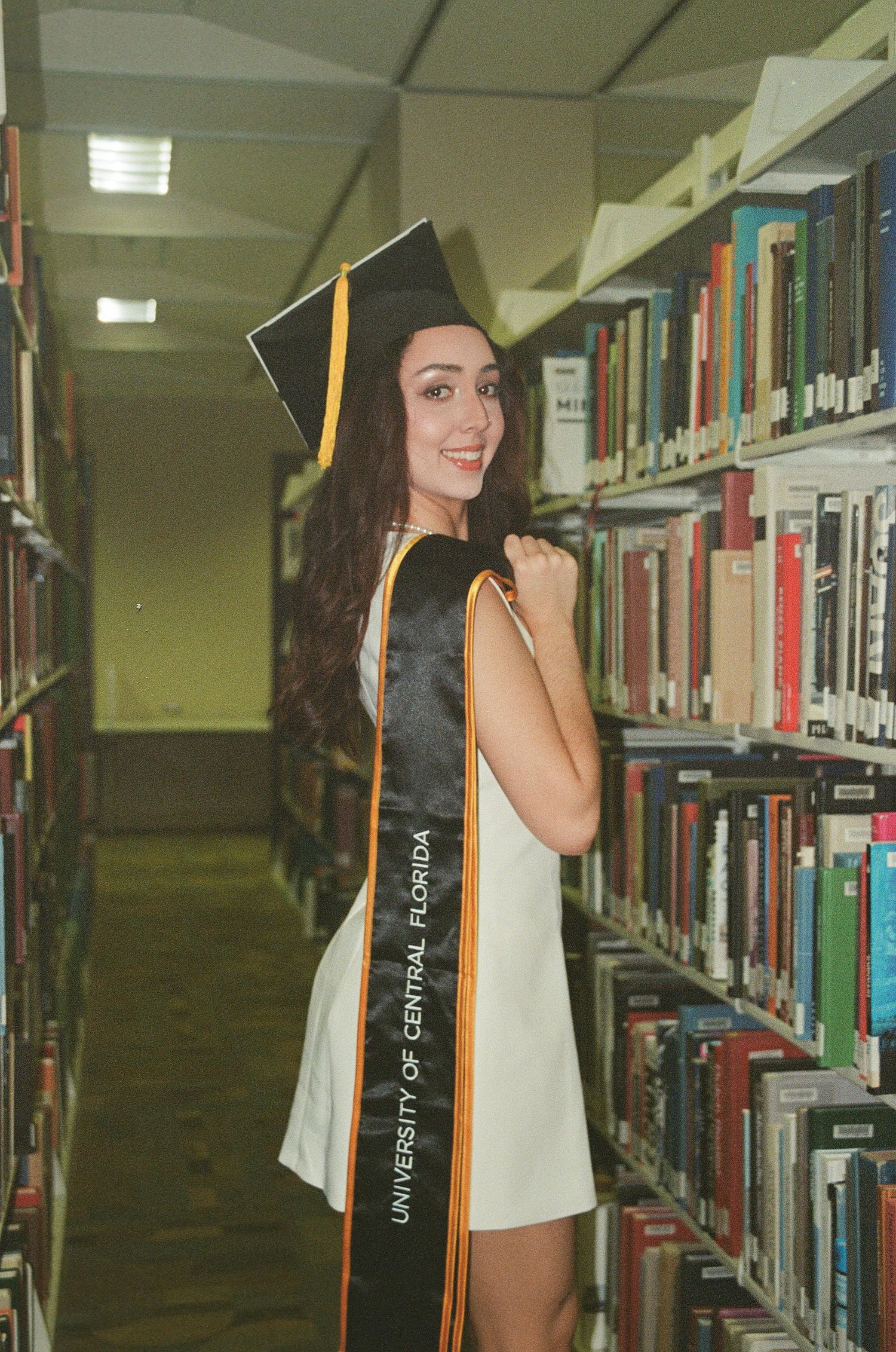 Smiling female UCF graduate in cap and gown poses beside library bookshelves.