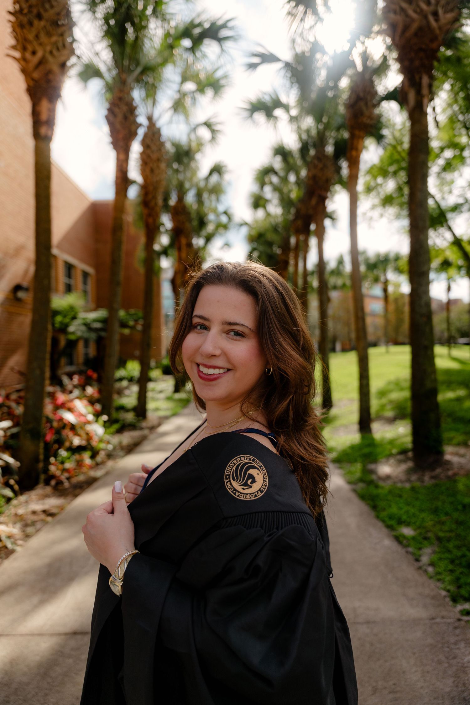 Smiling female graduate in black graduation gown with university seal posing on palm tree lined campus walkway.