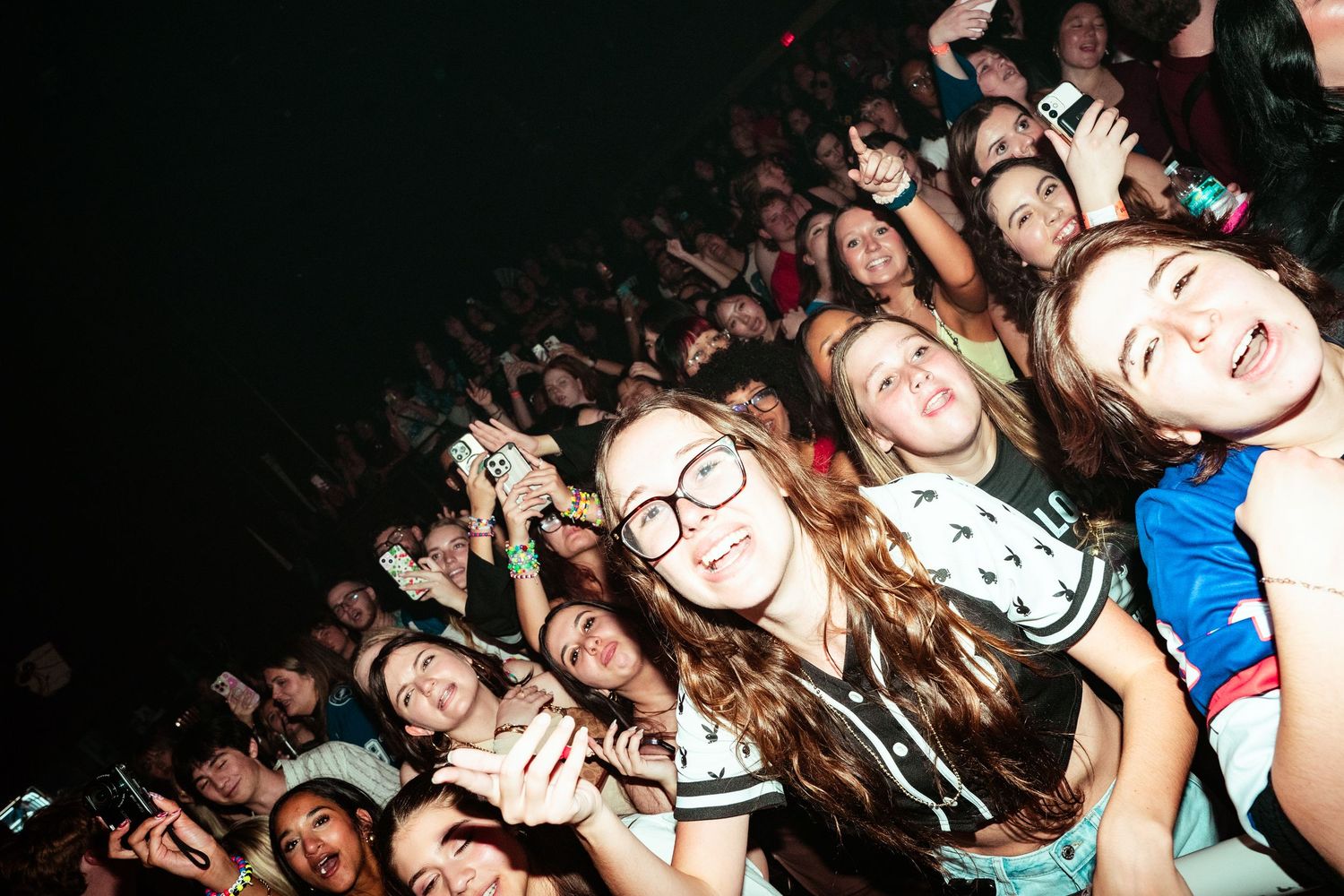 Enthusiastic crowd of young fans smiling and cheering at the front row of a live concert.