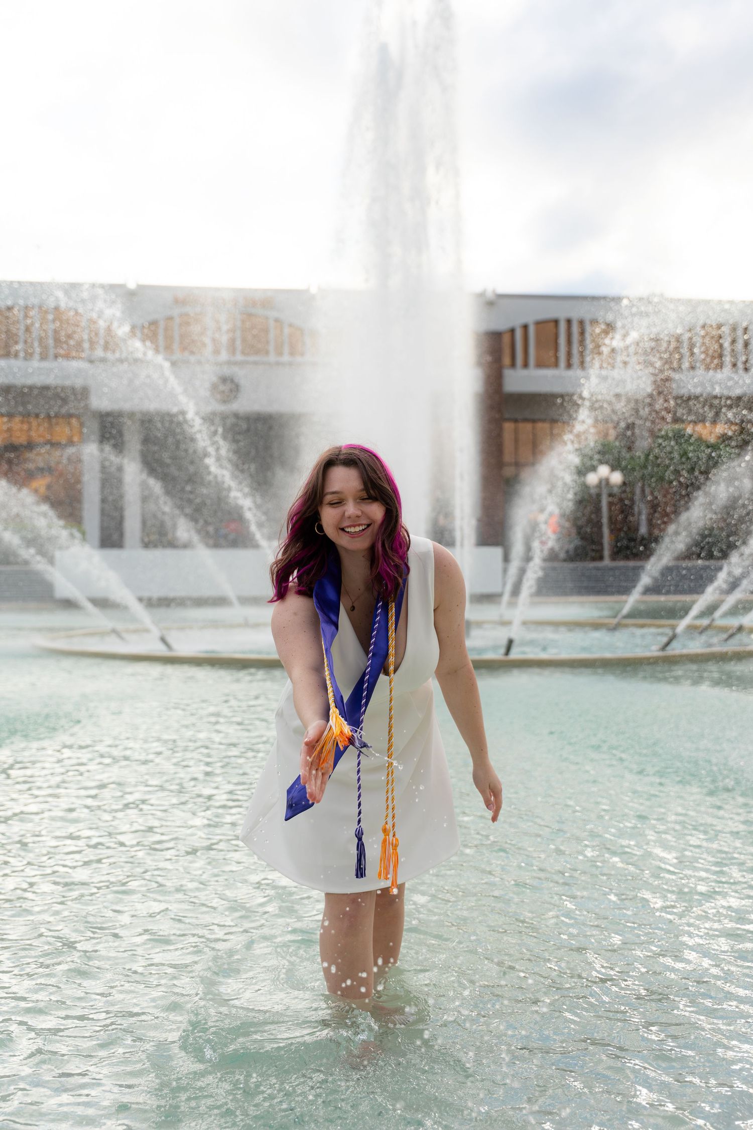 Smiling graduate in white dress with honor cords wades in UCF campus fountain.