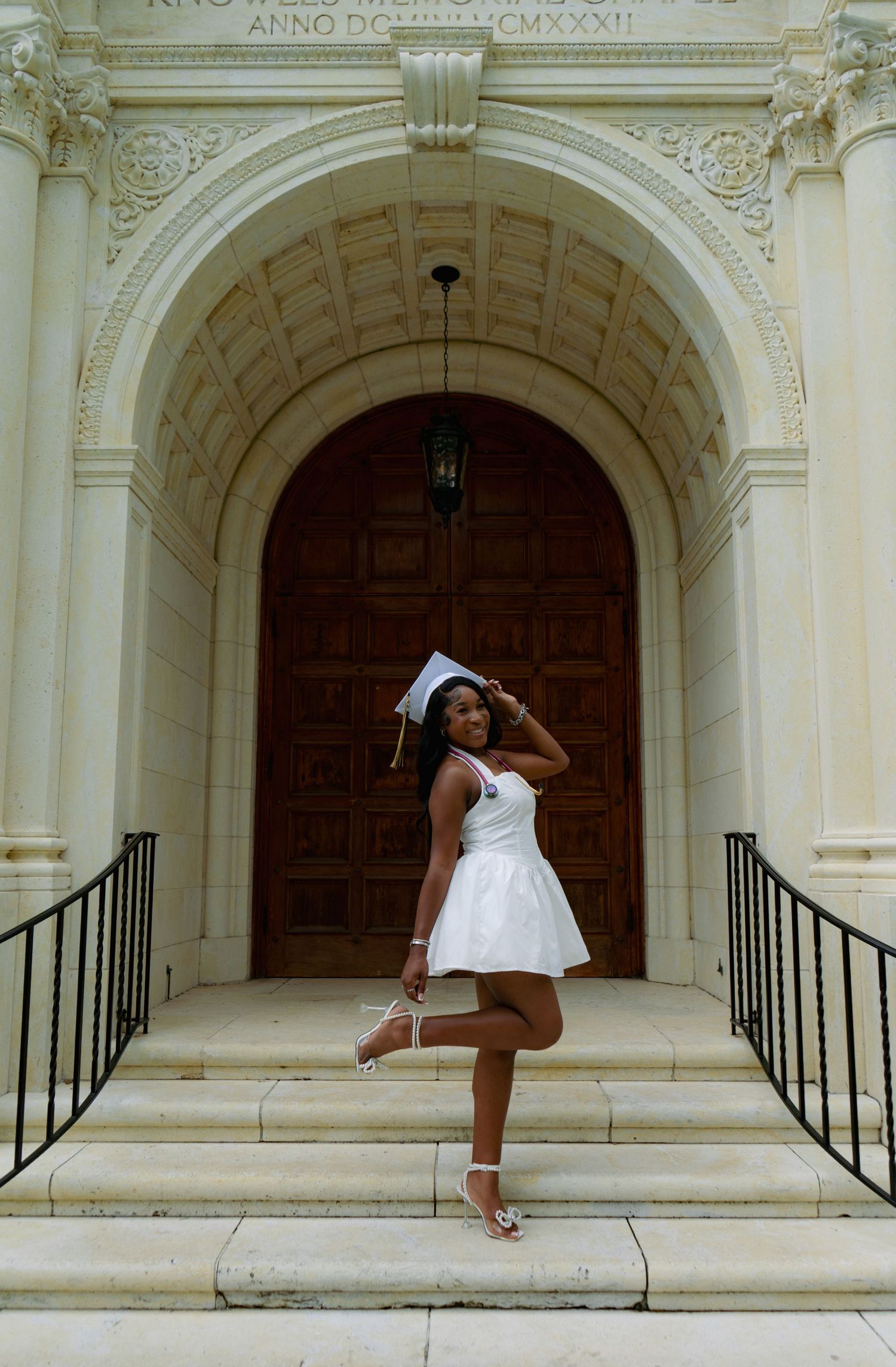 Graduate in white dress and cap poses on steps of a grand classical building with ornate archway.