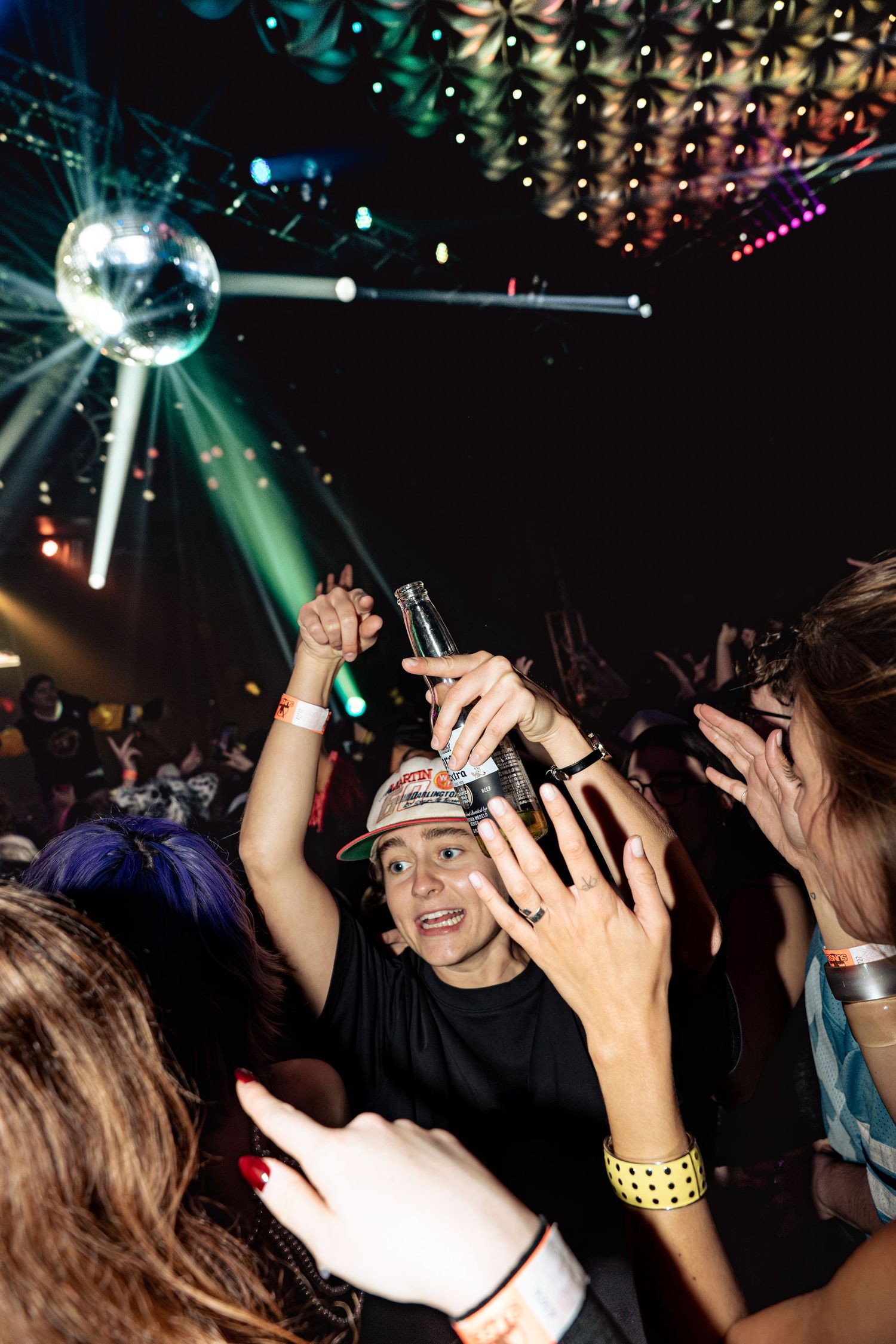 Person dancing in a nightclub crowd holding a bottle under a disco ball with colorful stage lights.