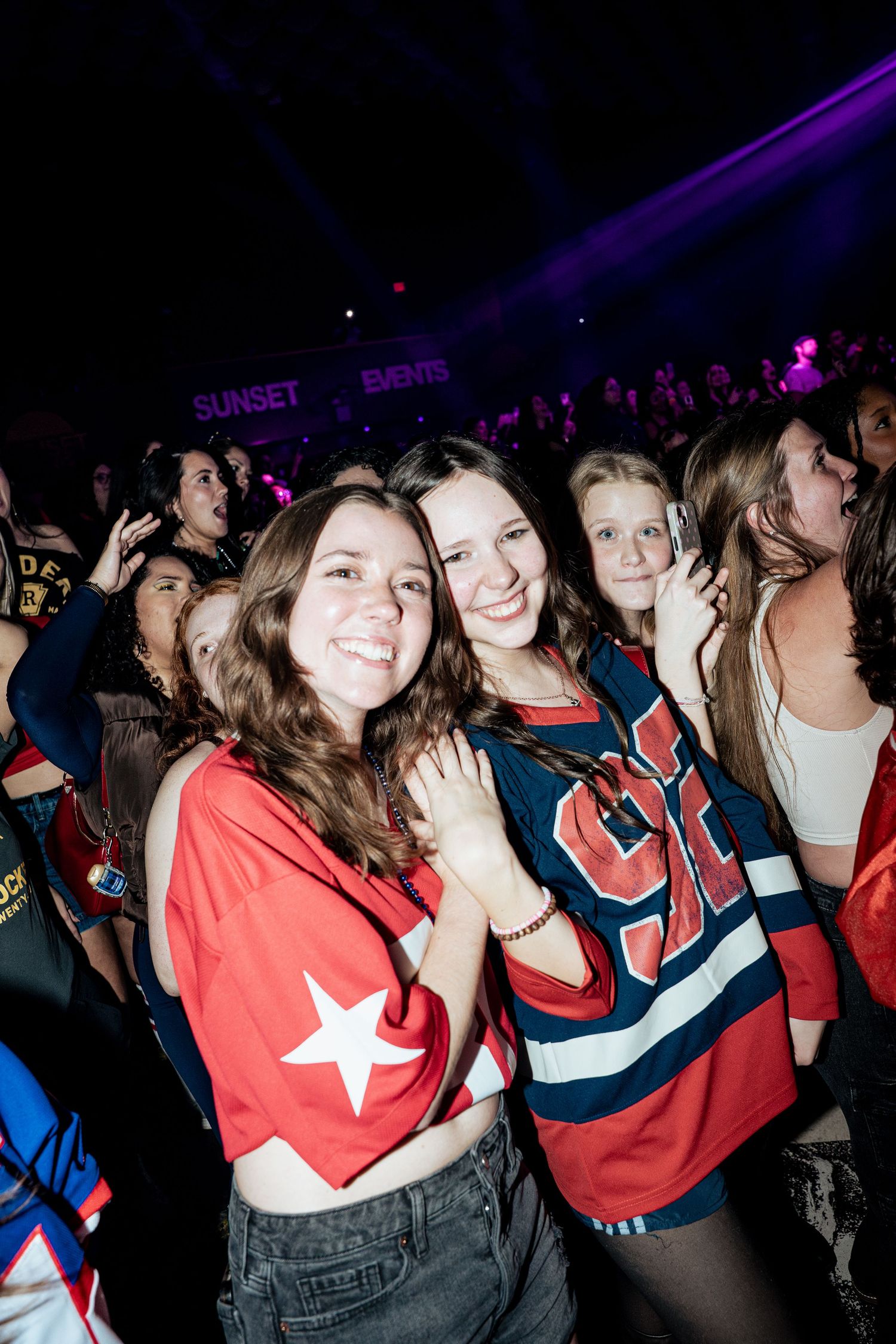 Two smiling young women in sports jerseys pose together at a lively concert venue with purple lights.