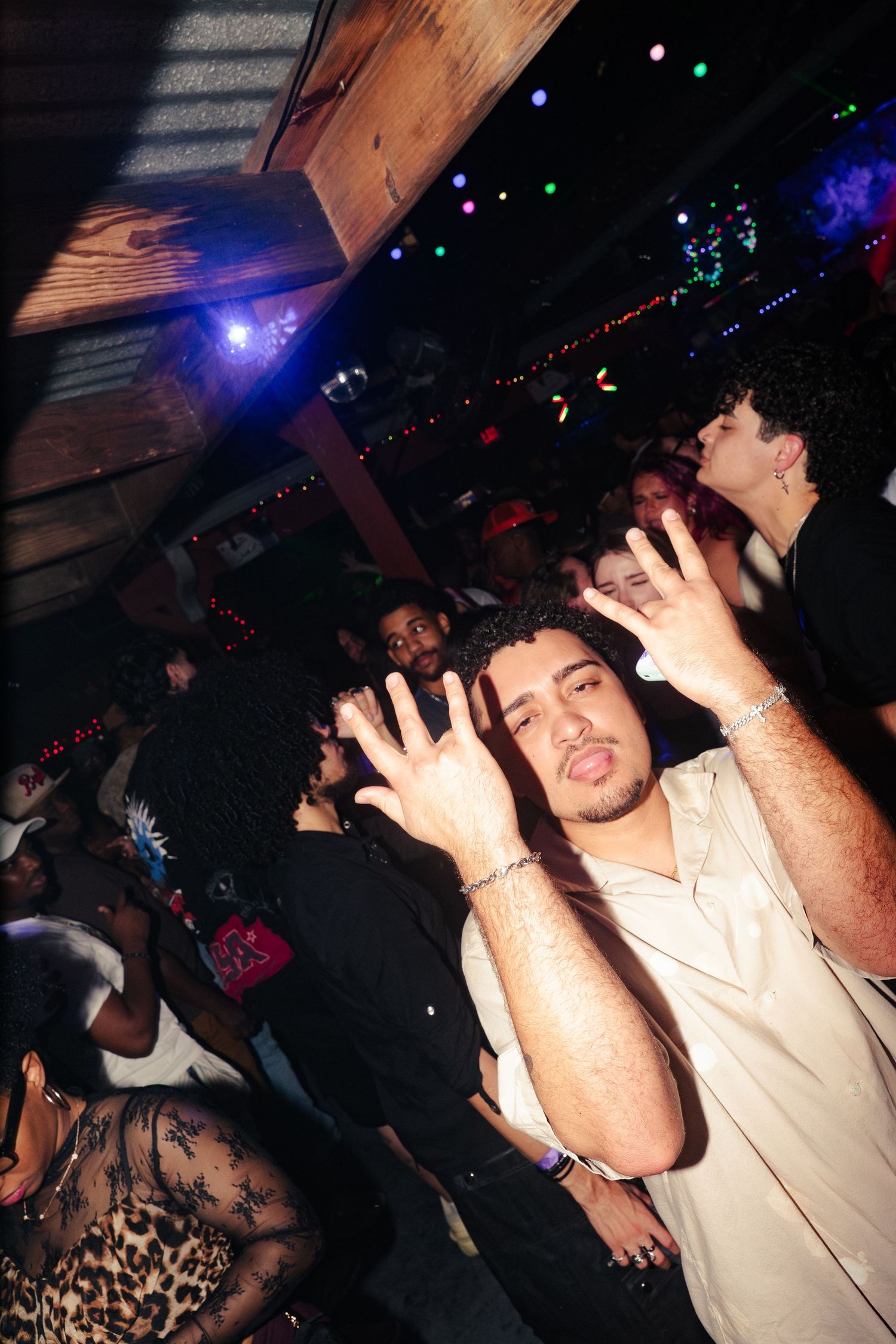 Young man posing with hands raised at a crowded nightclub party with colorful lights in the background.
