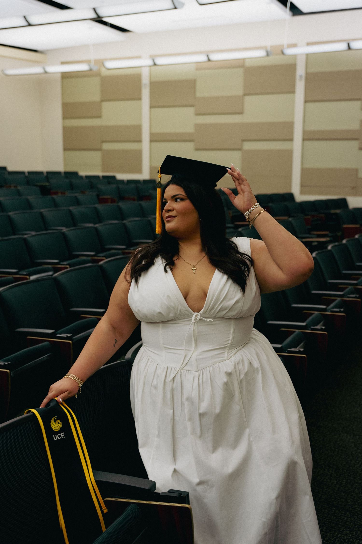 Graduate in white dress adjusts black cap while holding UCF stole in an empty auditorium.