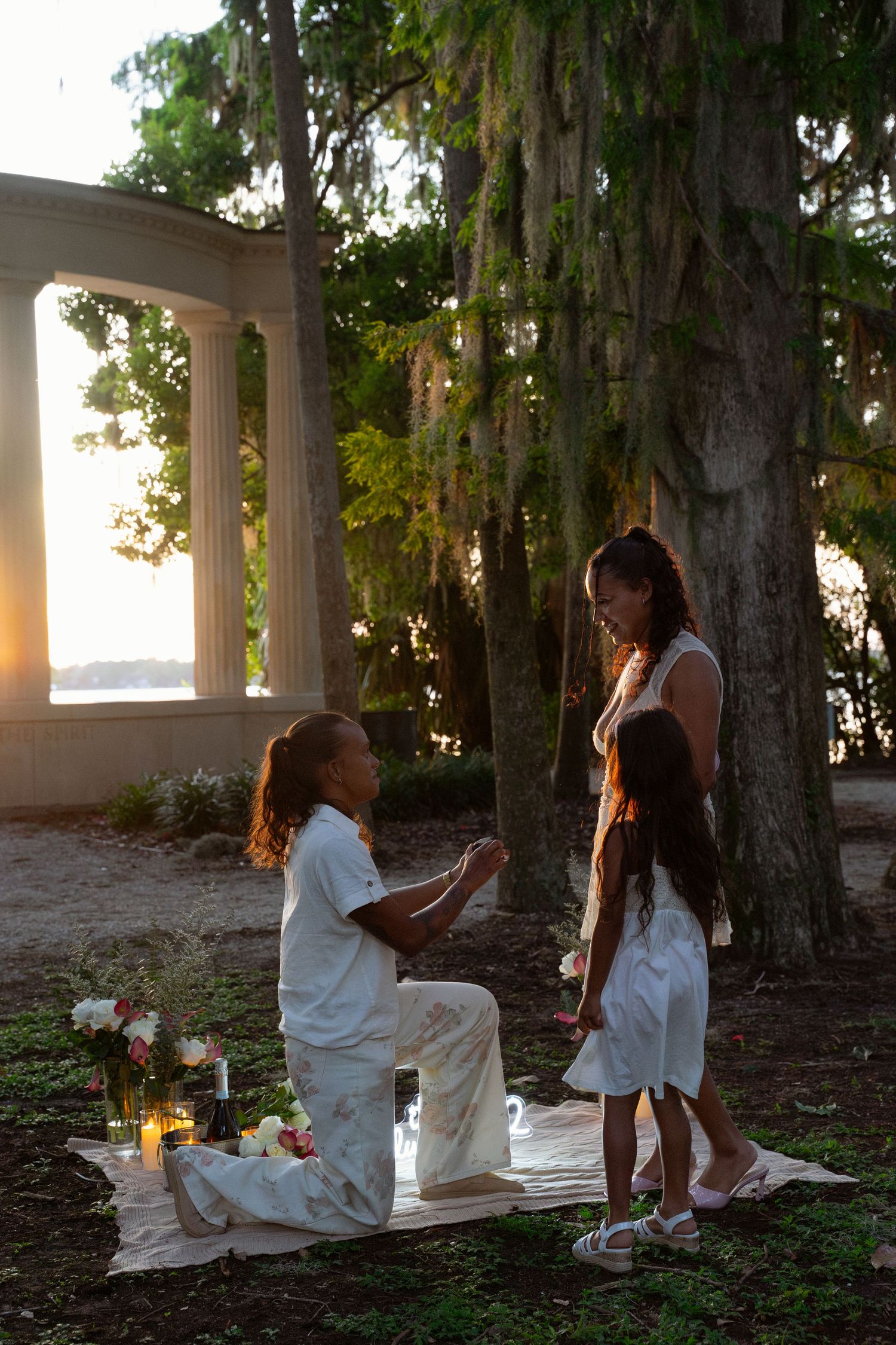 Romantic outdoor marriage proposal at sunset near moss-draped trees, man kneeling before woman with candles and flowers.