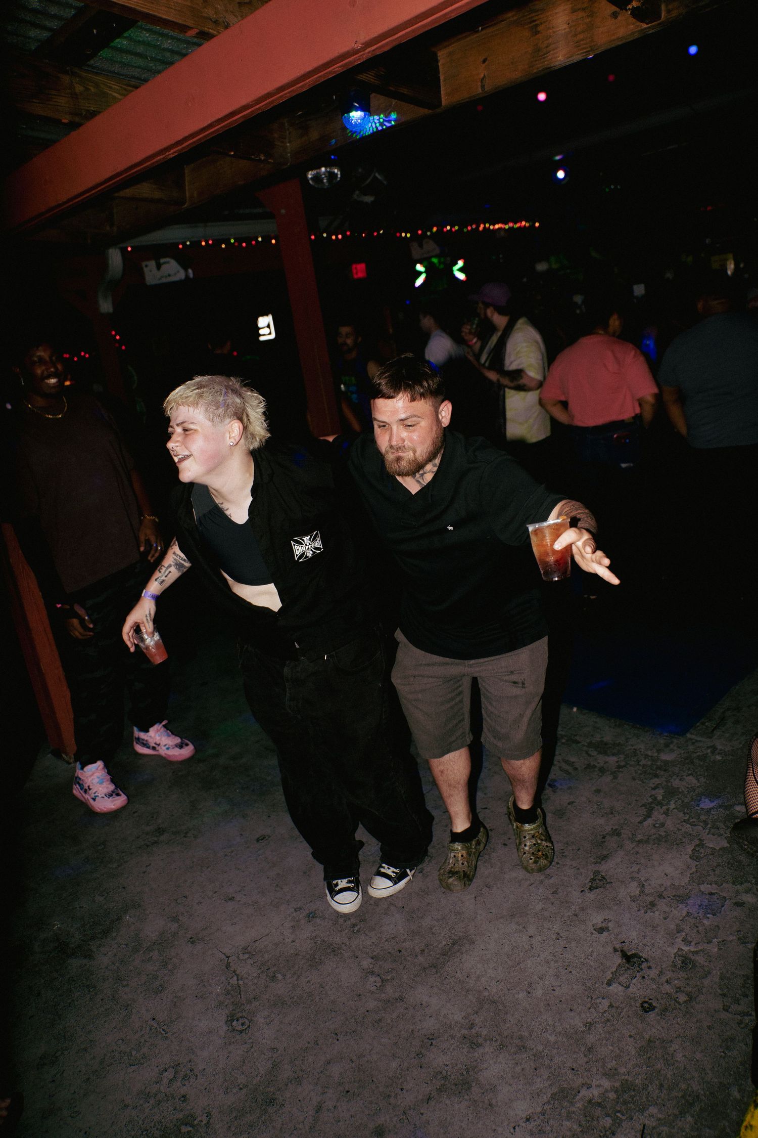 Two people dancing together at a dimly lit nightclub, one holding a drink while others socialize in the background.