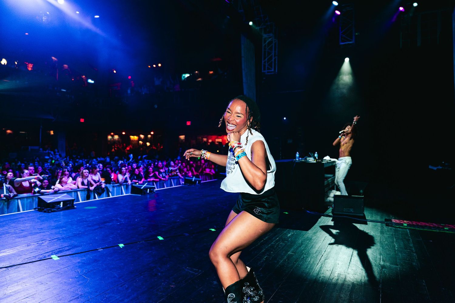 A woman performs energetically on stage at a packed concert venue with colorful purple and red lighting.