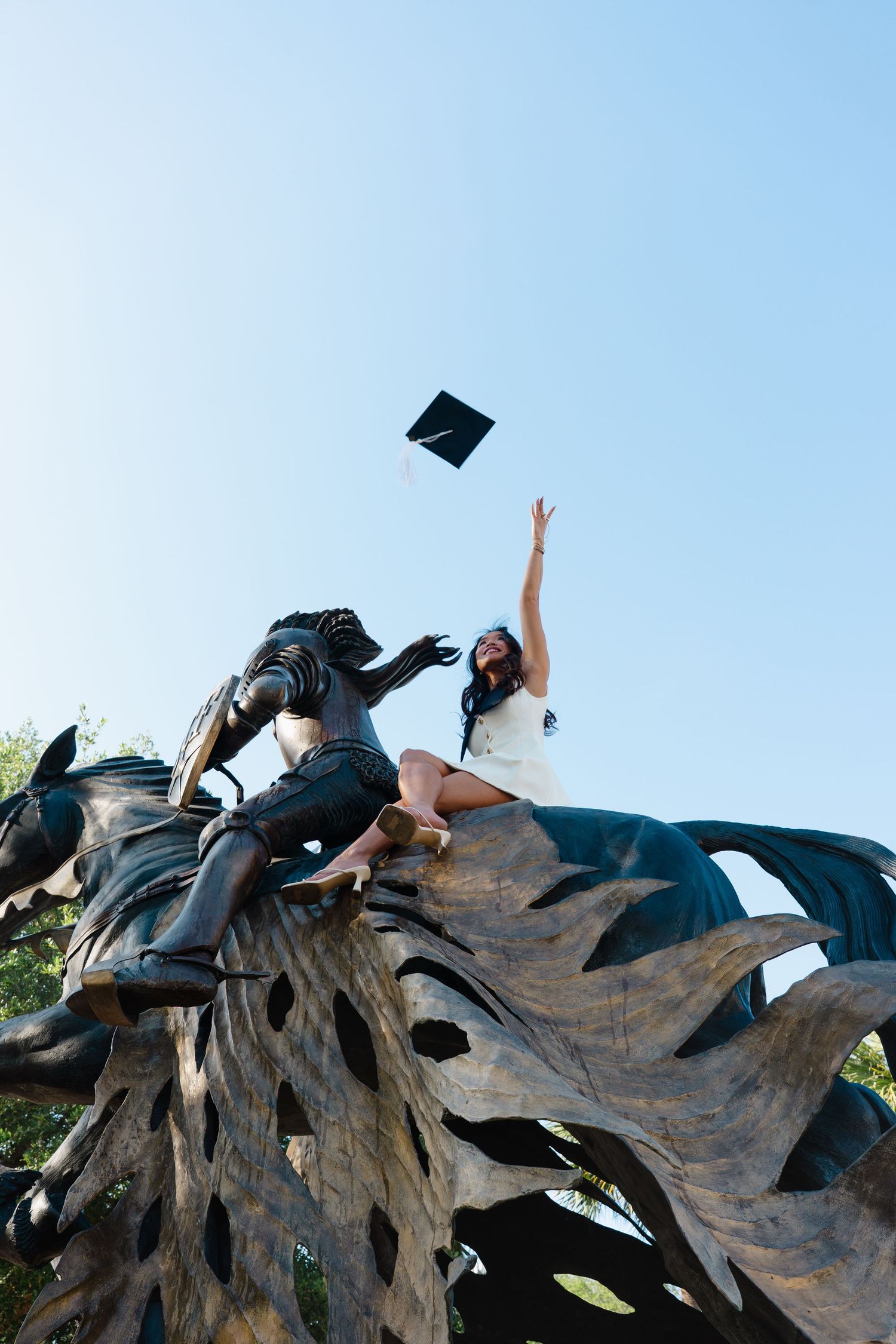 Graduate in white dress tosses cap from bronze statue base against clear blue sky.