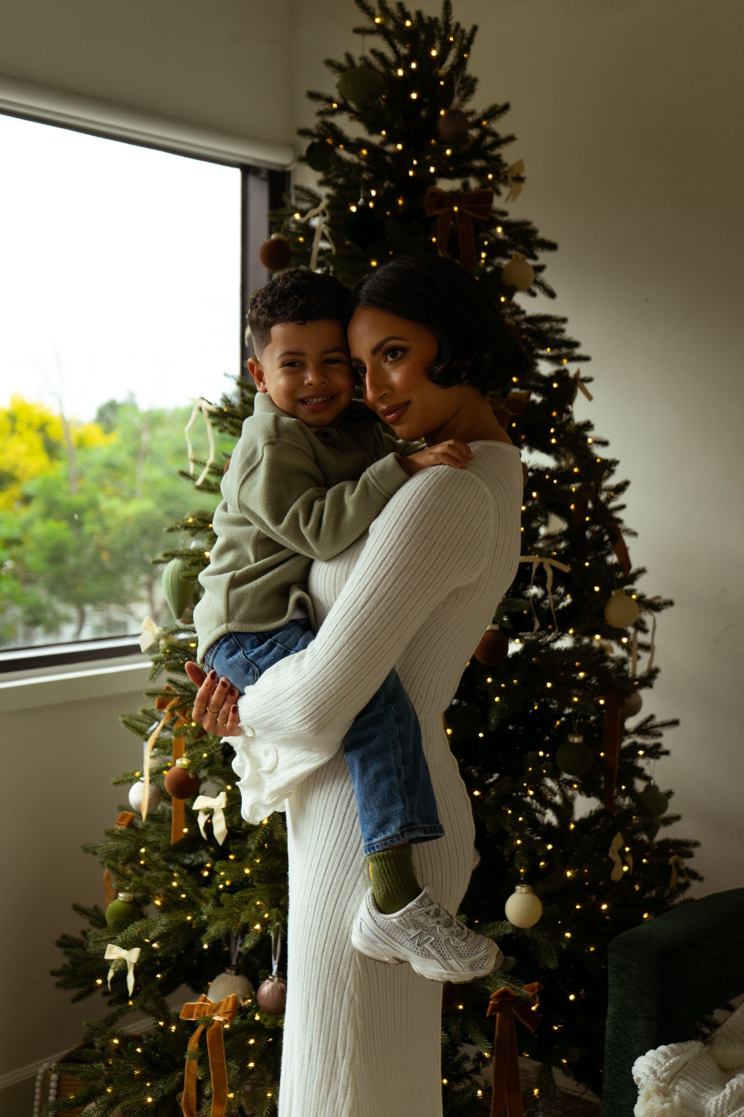 Smiling mother holding her young son in front of a beautifully decorated Christmas tree indoors.