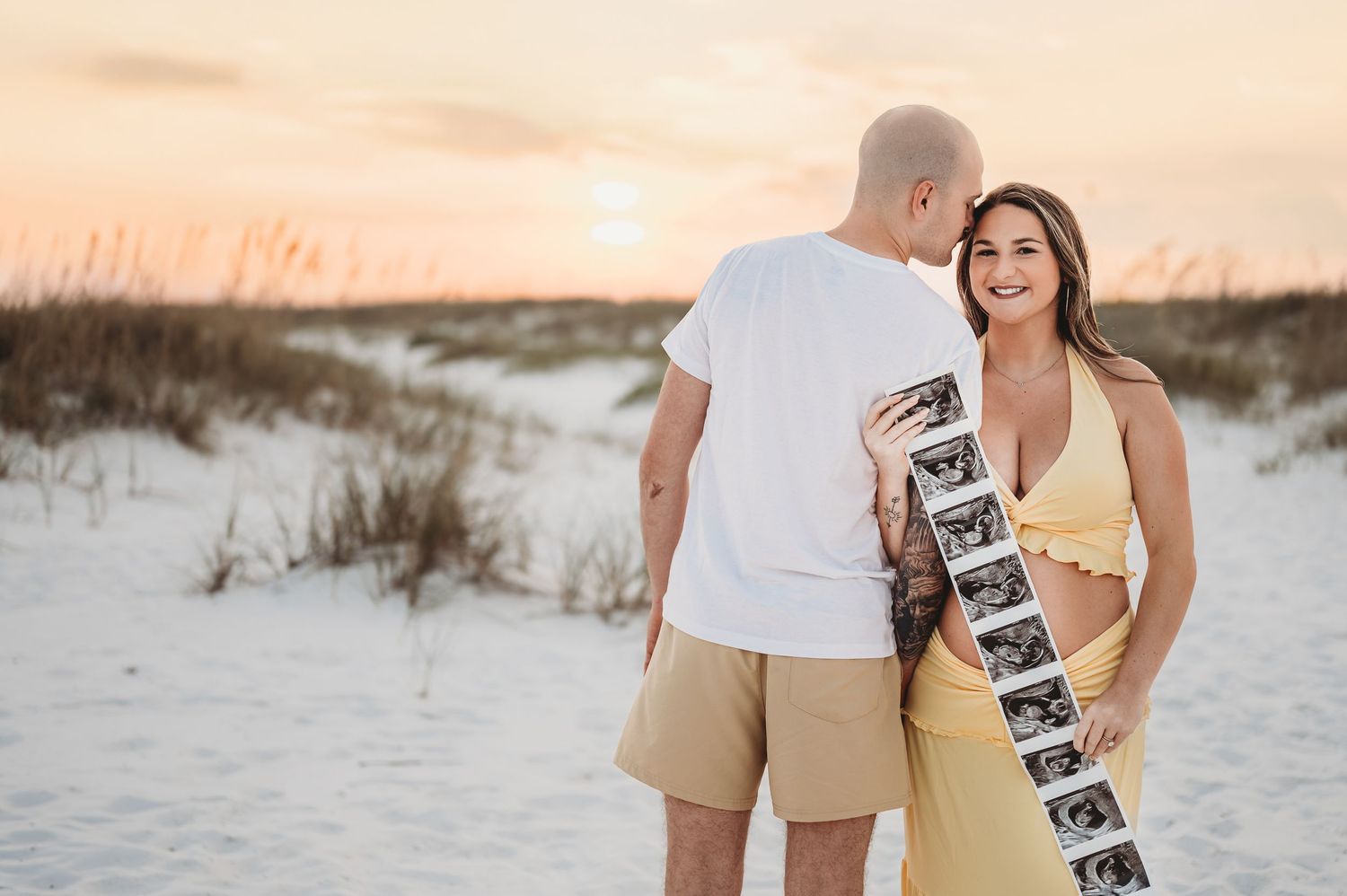Happy pregnant couple holding ultrasound photos on sandy beach dunes at golden sunset hour.