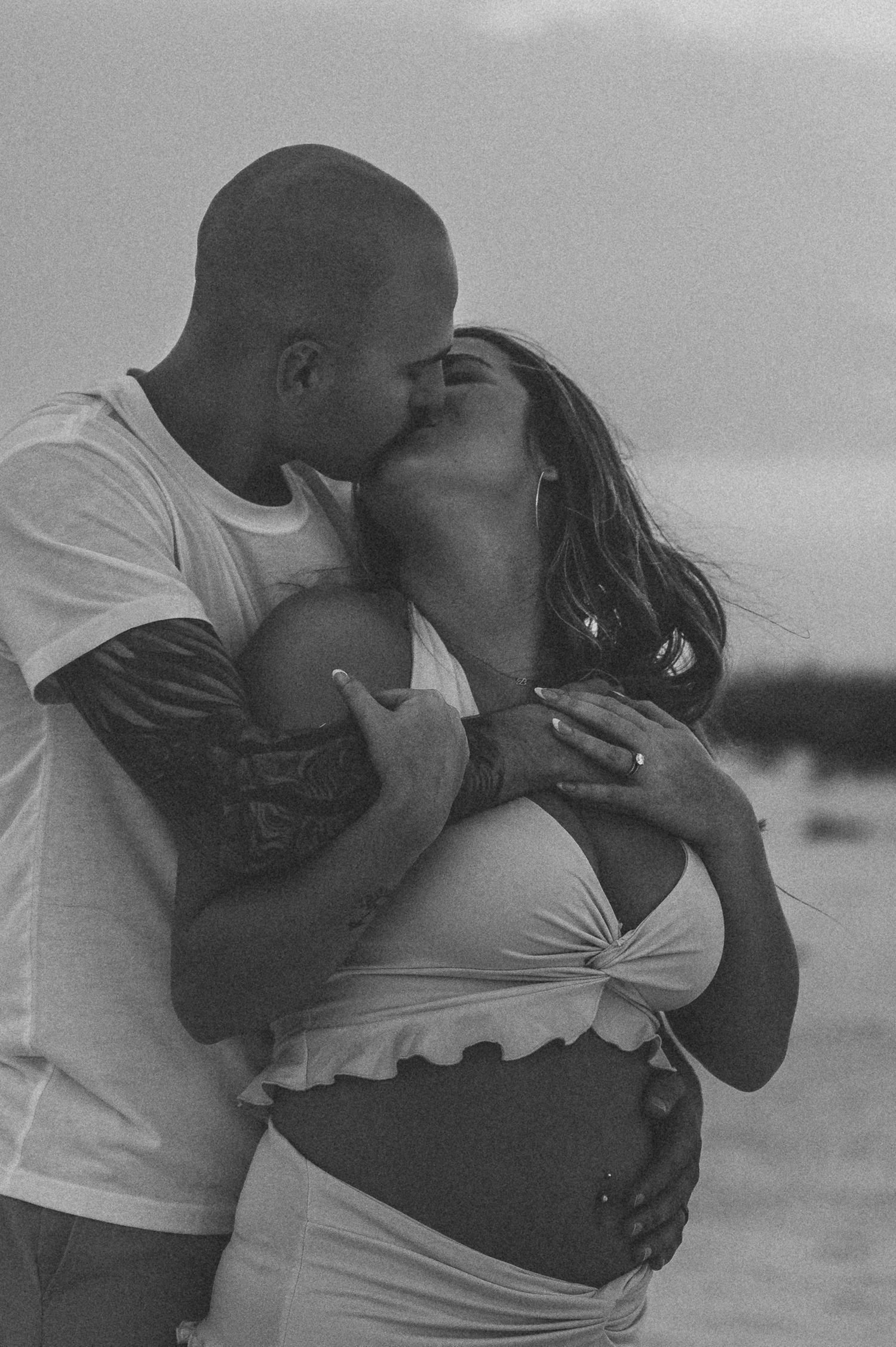 Couple sharing a kiss during maternity photoshoot on the beach, showing pregnant belly in black and white.