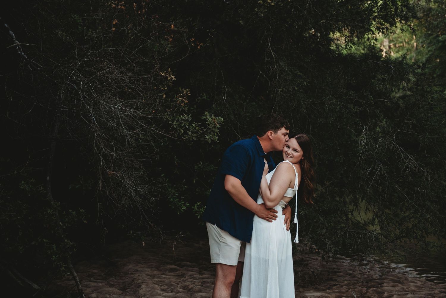 Couple sharing a tender kiss outdoors surrounded by lush dark green trees in natural light.