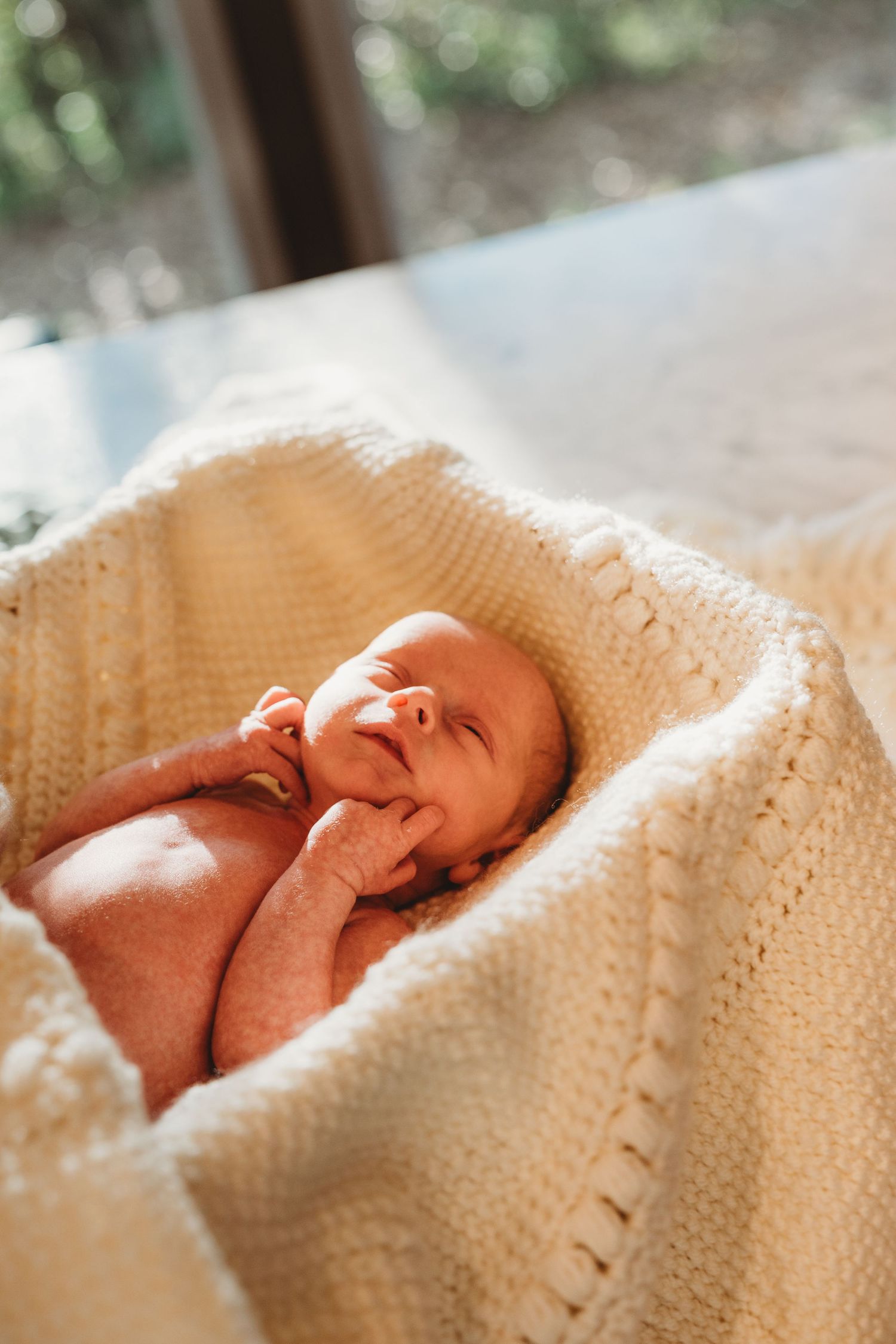 Smiling newborn baby wrapped in a cream knit blanket lying in a wicker bassinet in soft natural light.