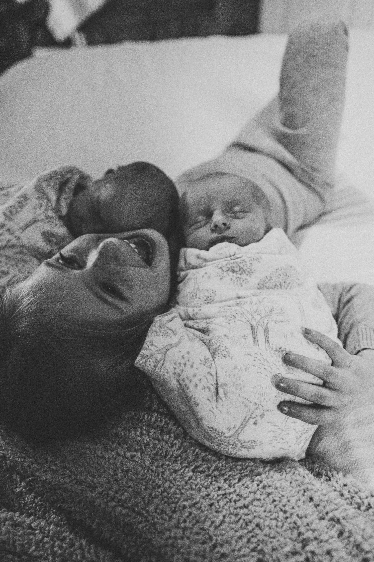 Black and white photo of a smiling mother lying down while cradling her sleeping newborn baby.