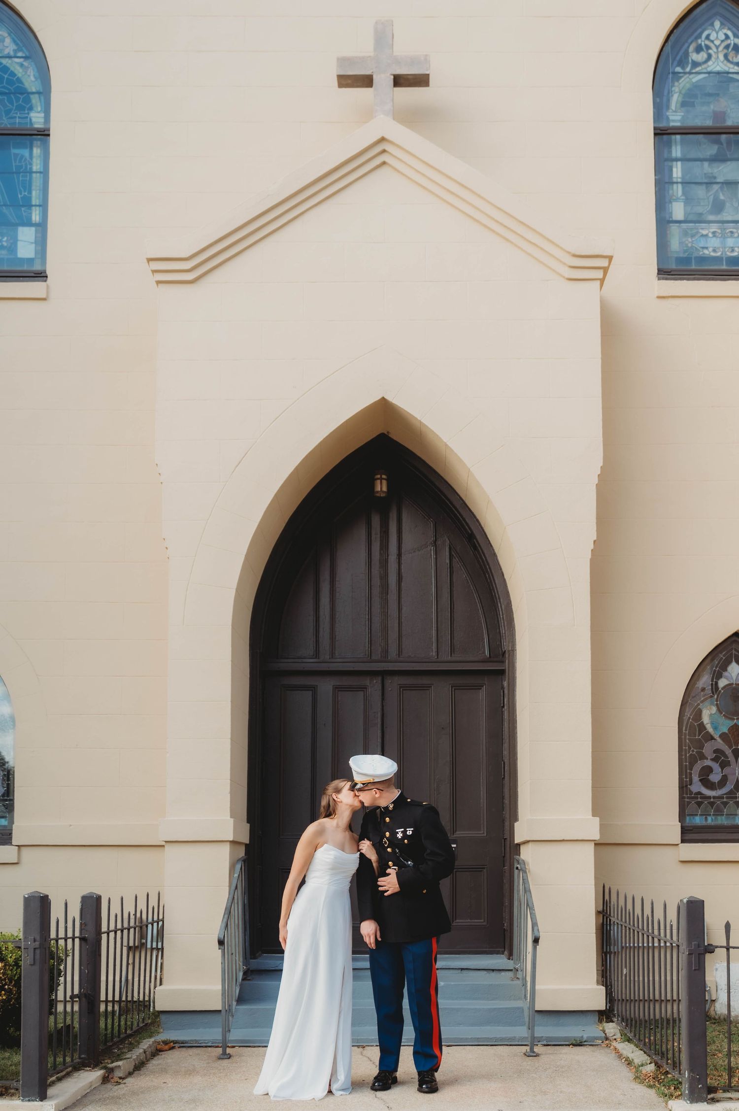 Marine groom kisses bride in white dress outside church arched wooden doors with cross.