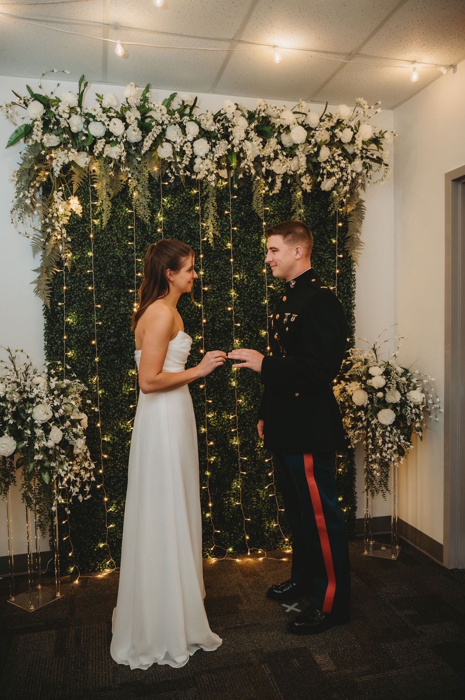 Couple exchanges rings at indoor ceremony before lush greenery wall with white florals and fairy lights.
