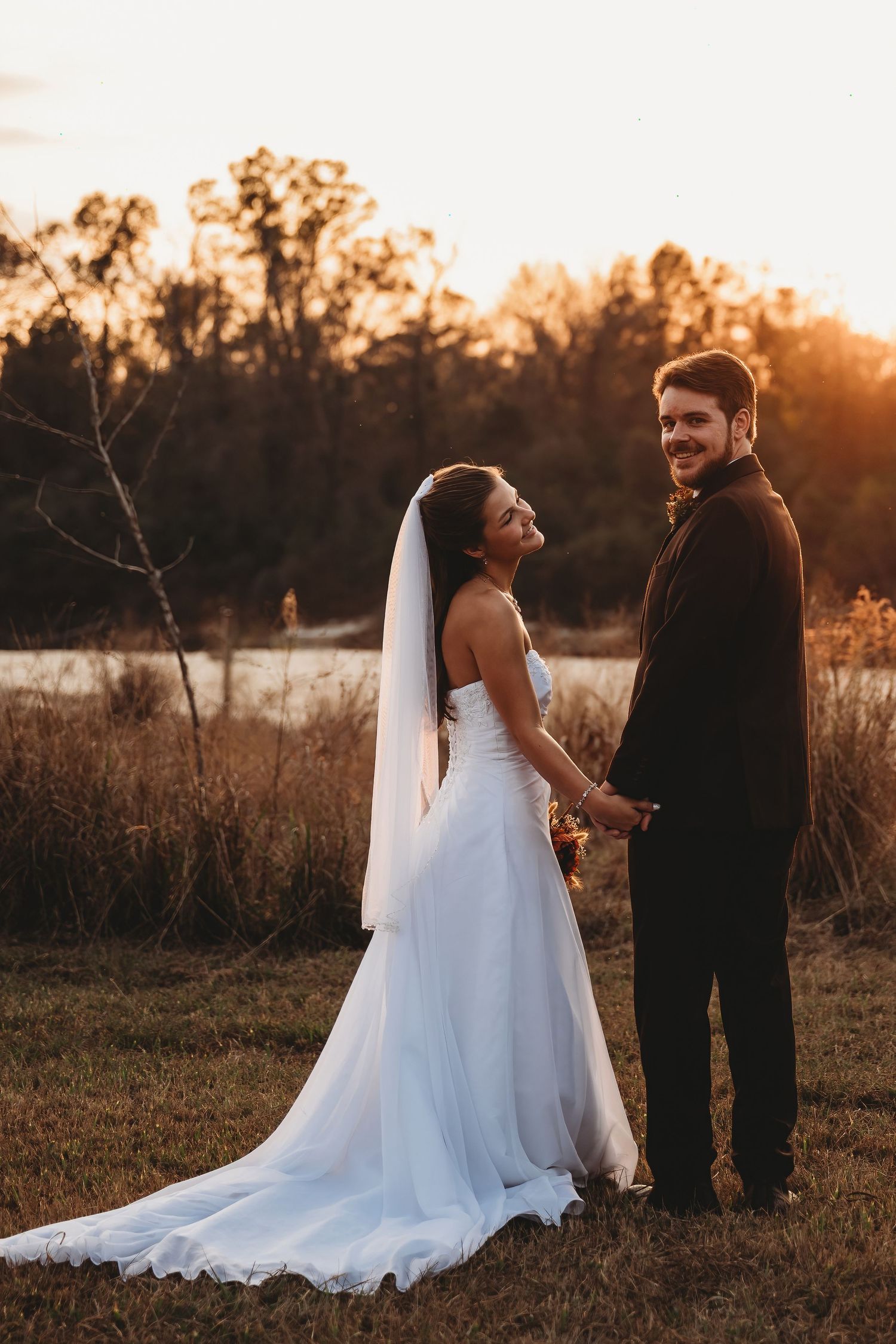 Bride and groom hold hands at sunset in a rustic outdoor setting, bride wearing a flowing white gown with veil.