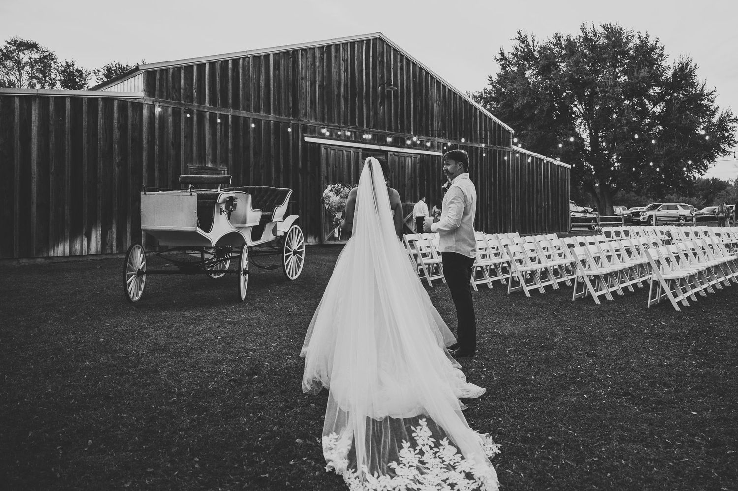 Black and white wedding photo of bride in flowing veil with groom near vintage carriage and barn venue.