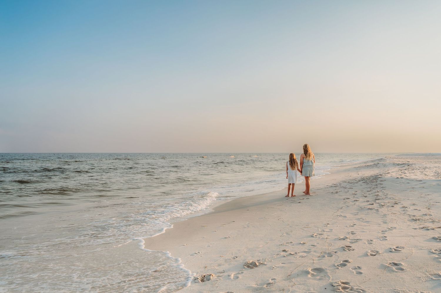Two children stand hand-in-hand on a white sandy beach at sunset, gazing at calm ocean waves.