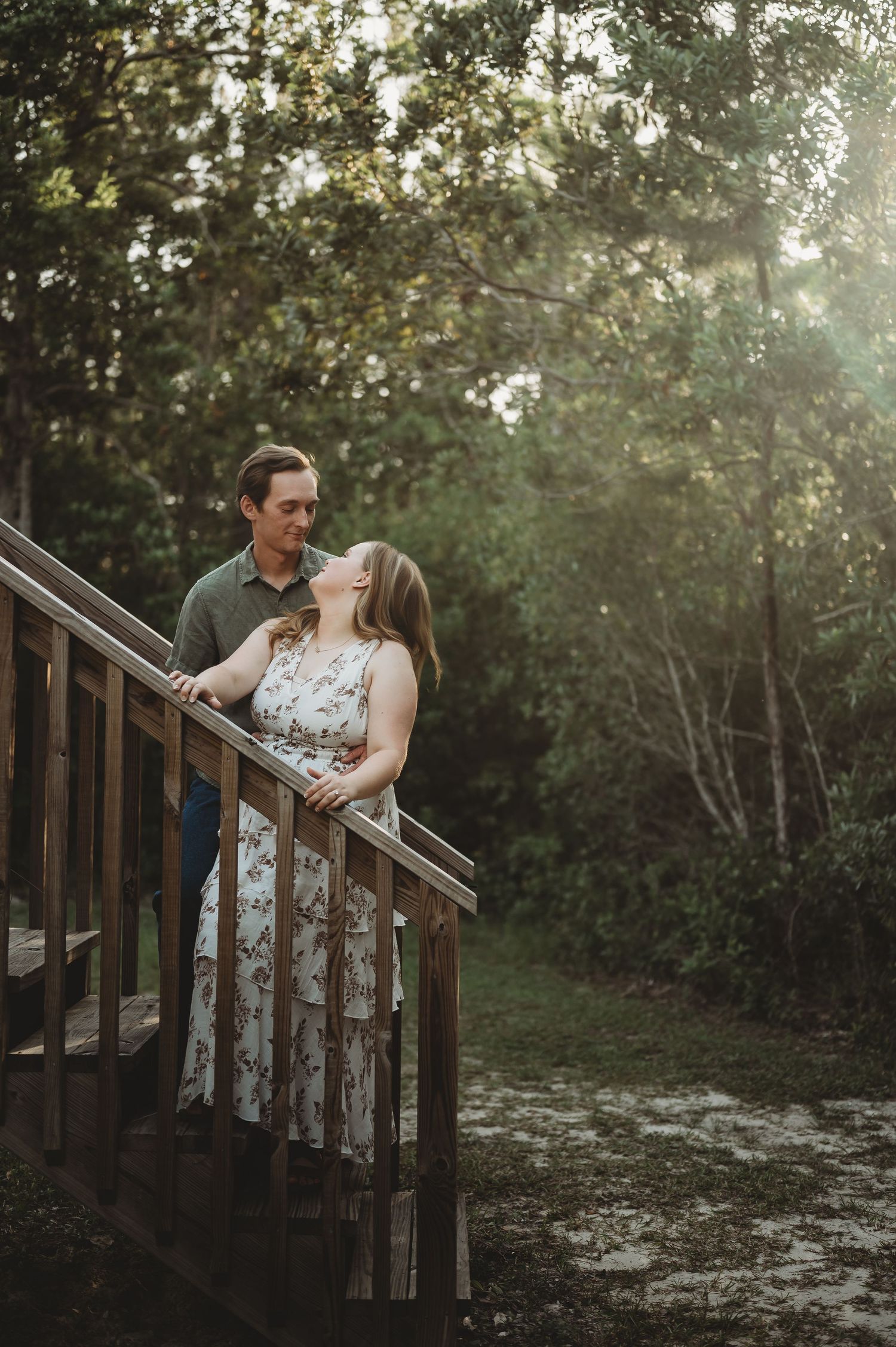 Couple sharing a romantic moment on a wooden bridge surrounded by lush green trees in golden hour light.