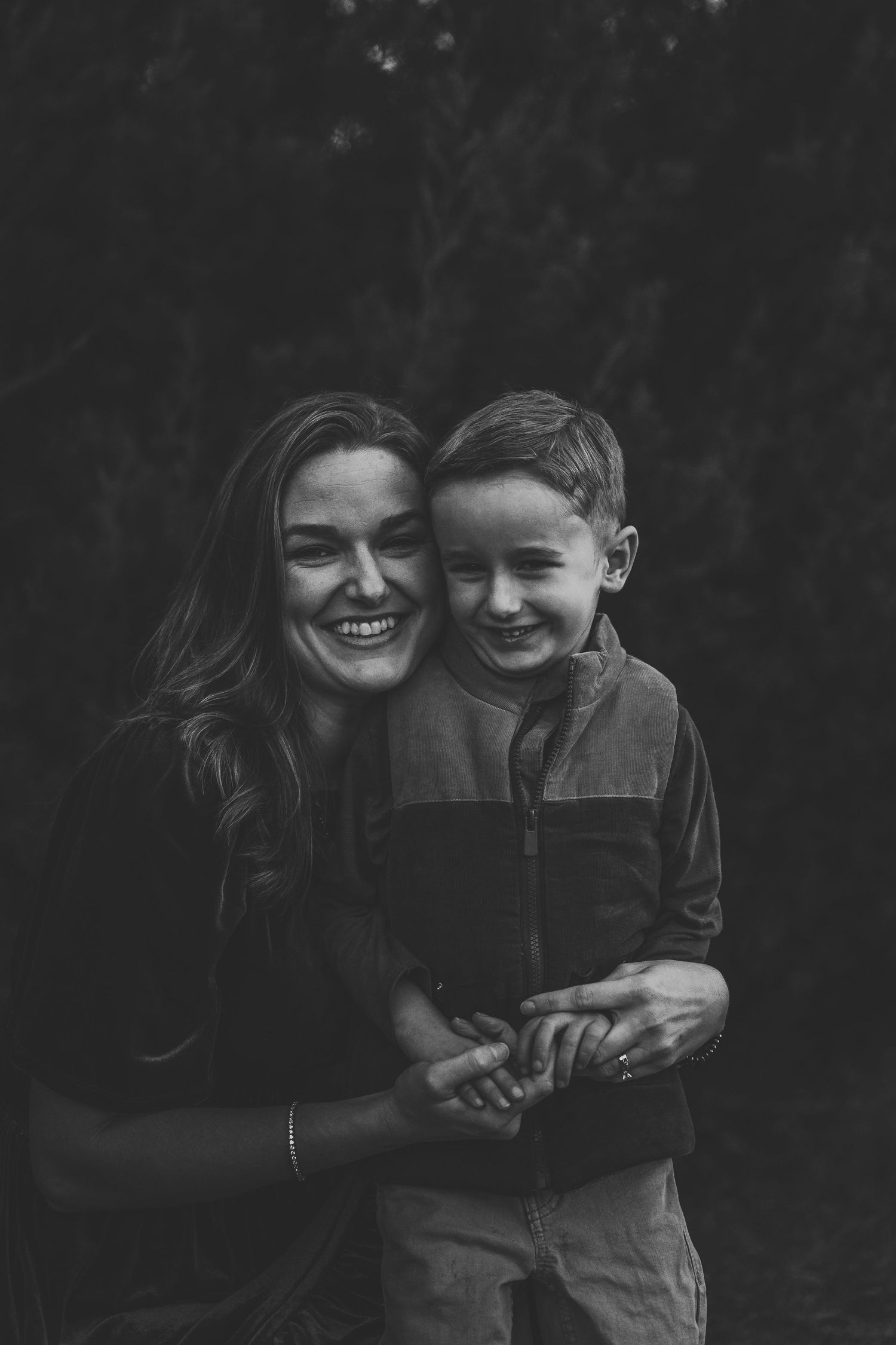Black and white photo of a smiling woman hugging a young boy outdoors, both laughing joyfully.