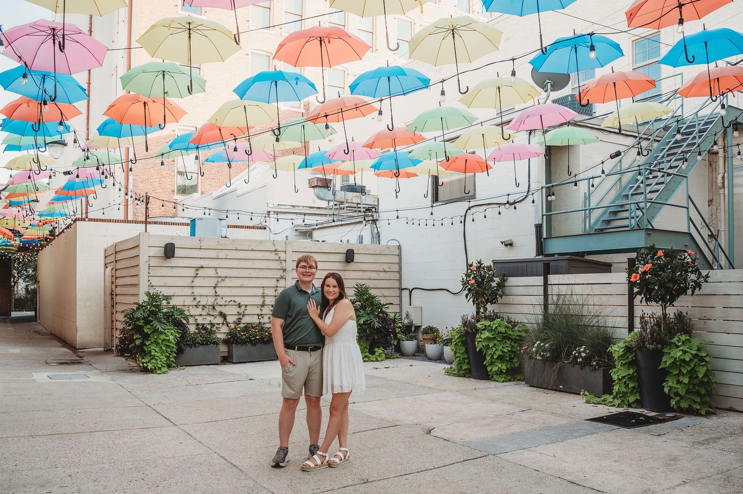 Couple smiling under colorful hanging umbrellas in a vibrant outdoor courtyard with potted plants and string lights.