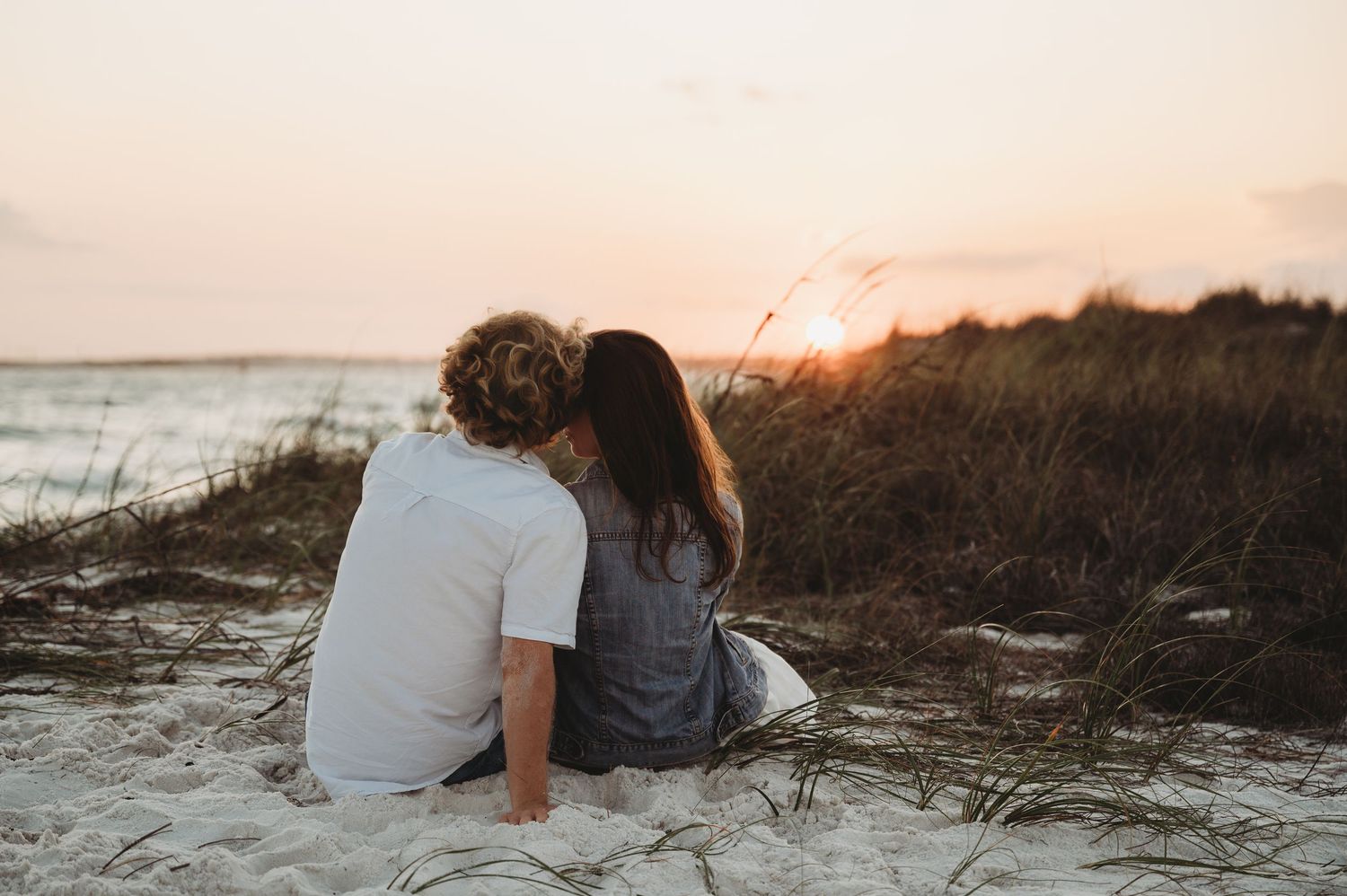 Couple sitting together on sandy beach watching romantic sunset over ocean dunes in warm golden light.