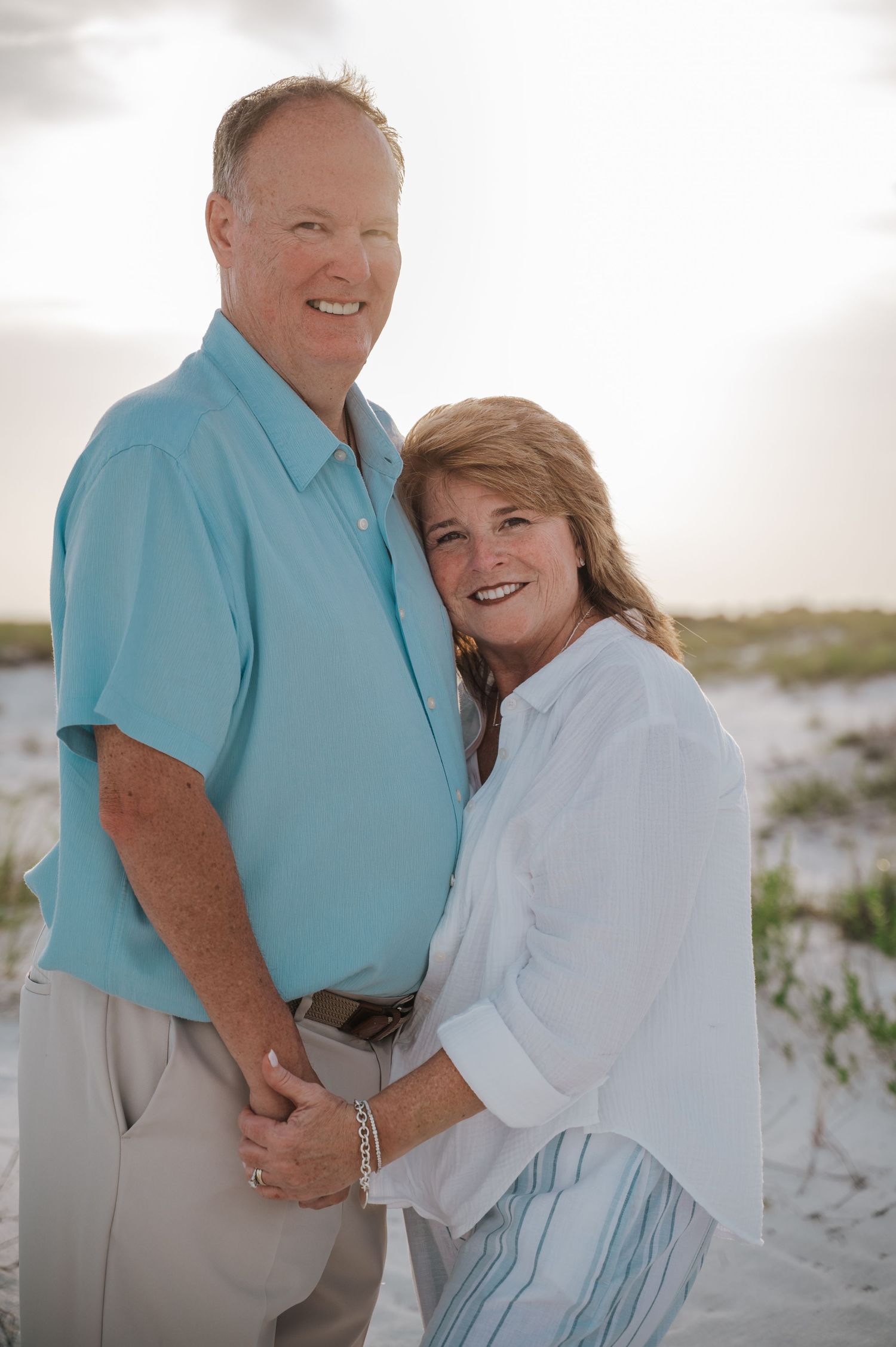 Smiling couple posing on sandy beach at sunset, man in teal shirt and woman in white blouse holding hands.
