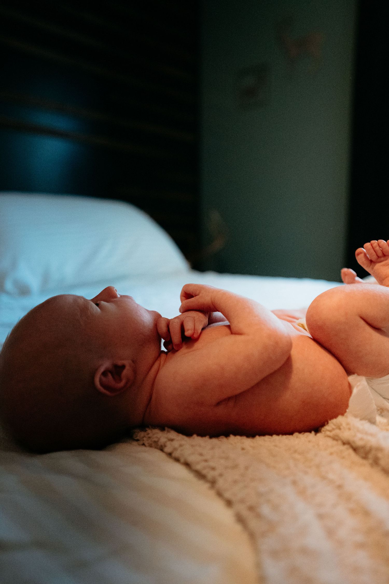 Newborn baby lying unswaddled on a fluffy blanket on a white bed in a softly lit room.