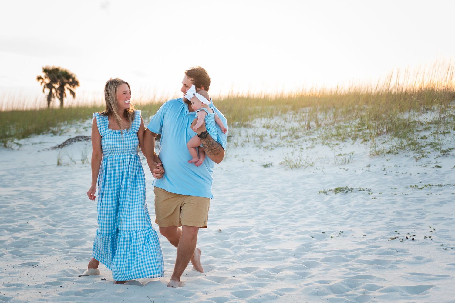 Young family in blue outfits walking on a sandy beach at sunset with their baby.