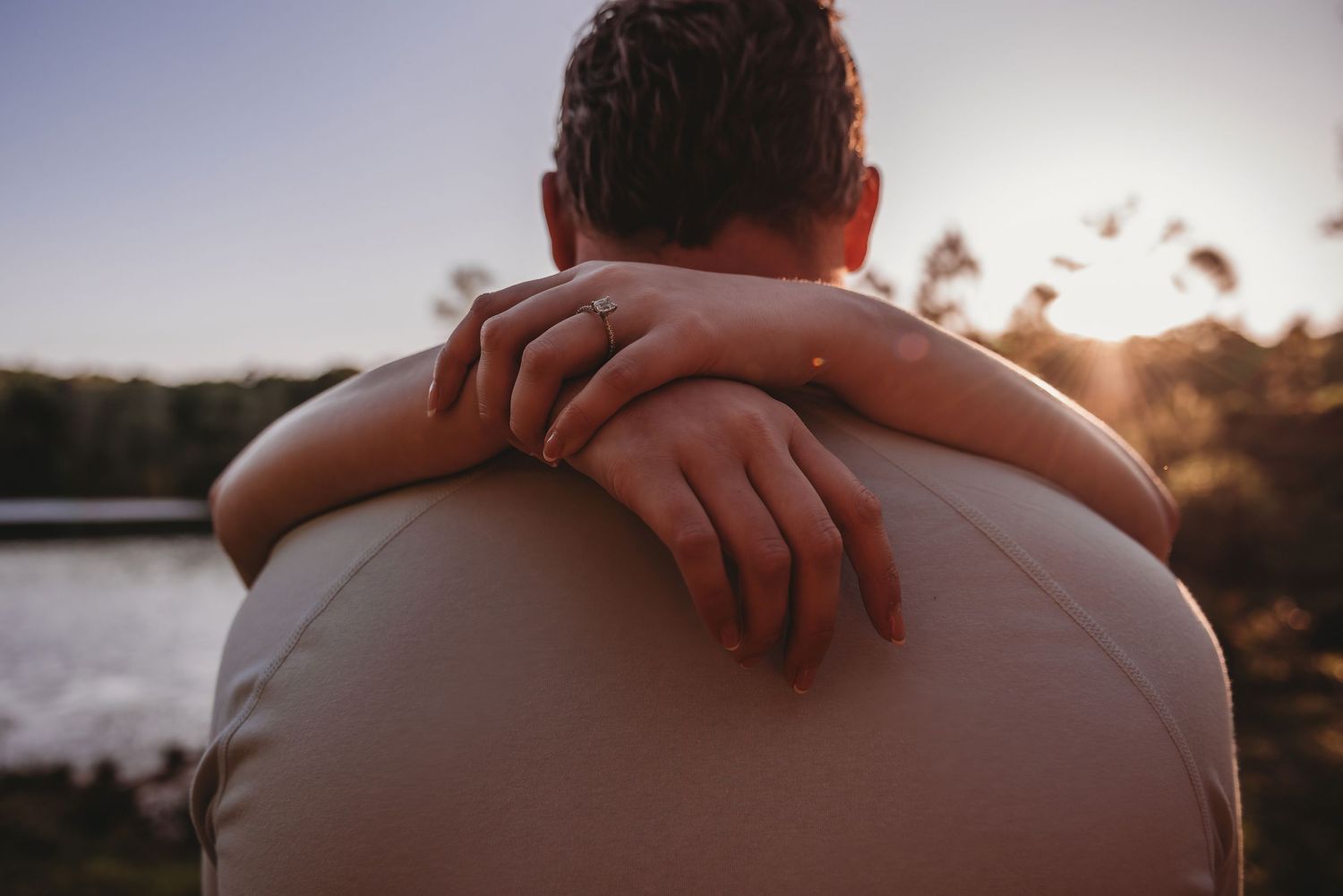 Close-up of a woman's hands with engagement ring wrapped around her partner's shoulders at sunset.