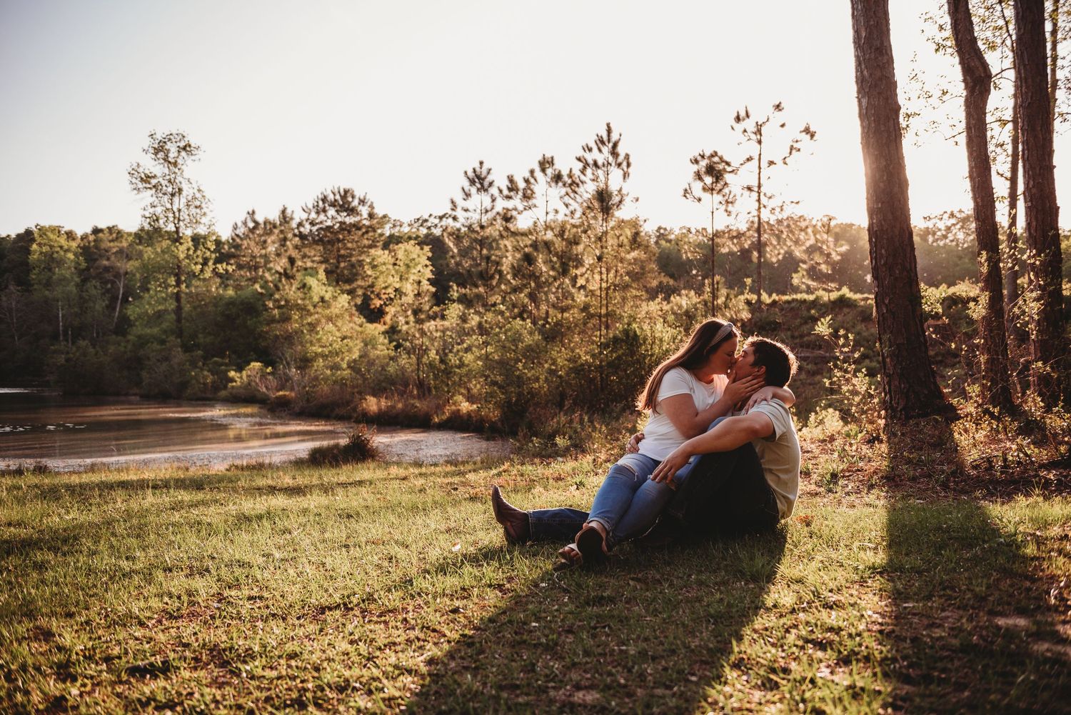 Couple sitting closely together on sunny grass near a lake, sharing a romantic kiss outdoors.