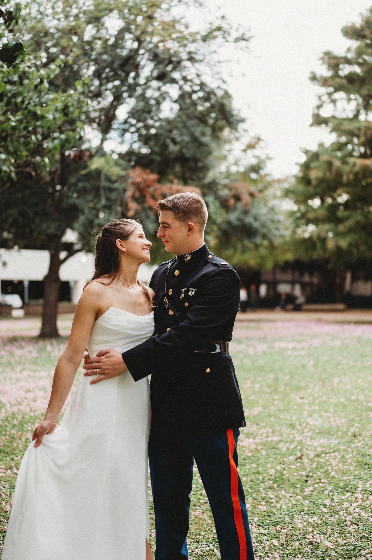 Marine groom in dress blues smiles at bride in white gown outdoors.