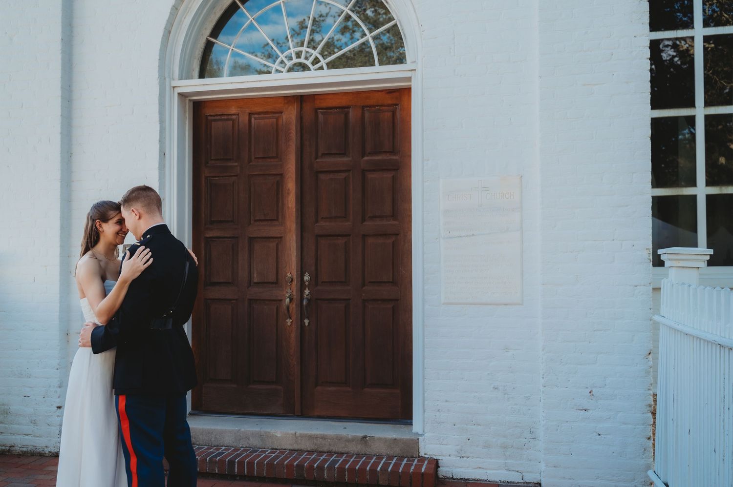 Bride and groom embracing before elegant wooden doors of white brick church on their wedding day.