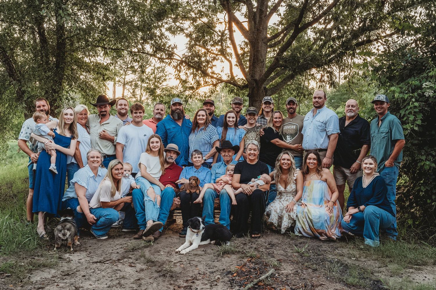 Large multigenerational family group portrait outdoors surrounded by lush green trees during golden hour.