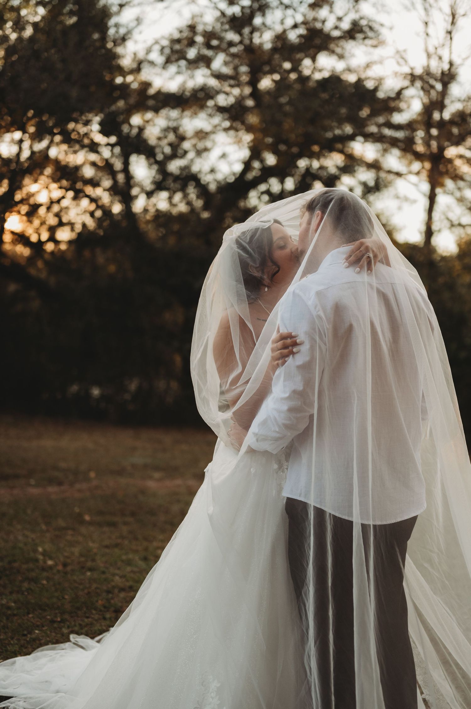 Bride and groom sharing a romantic kiss under a flowing wedding veil outdoors at golden hour.