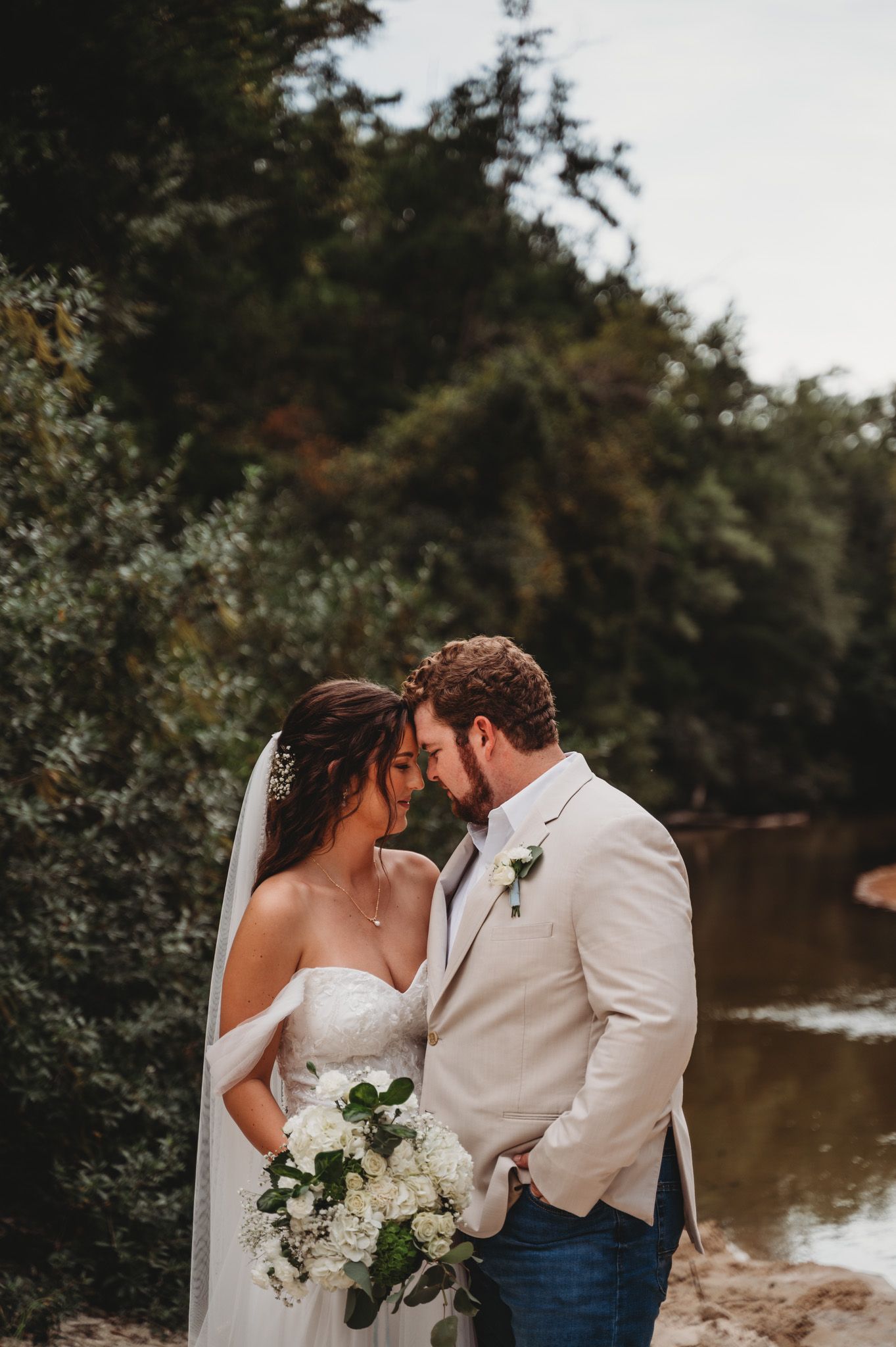Bride and groom touching foreheads romantically near a river, bride holding white hydrangea bouquet outdoors.