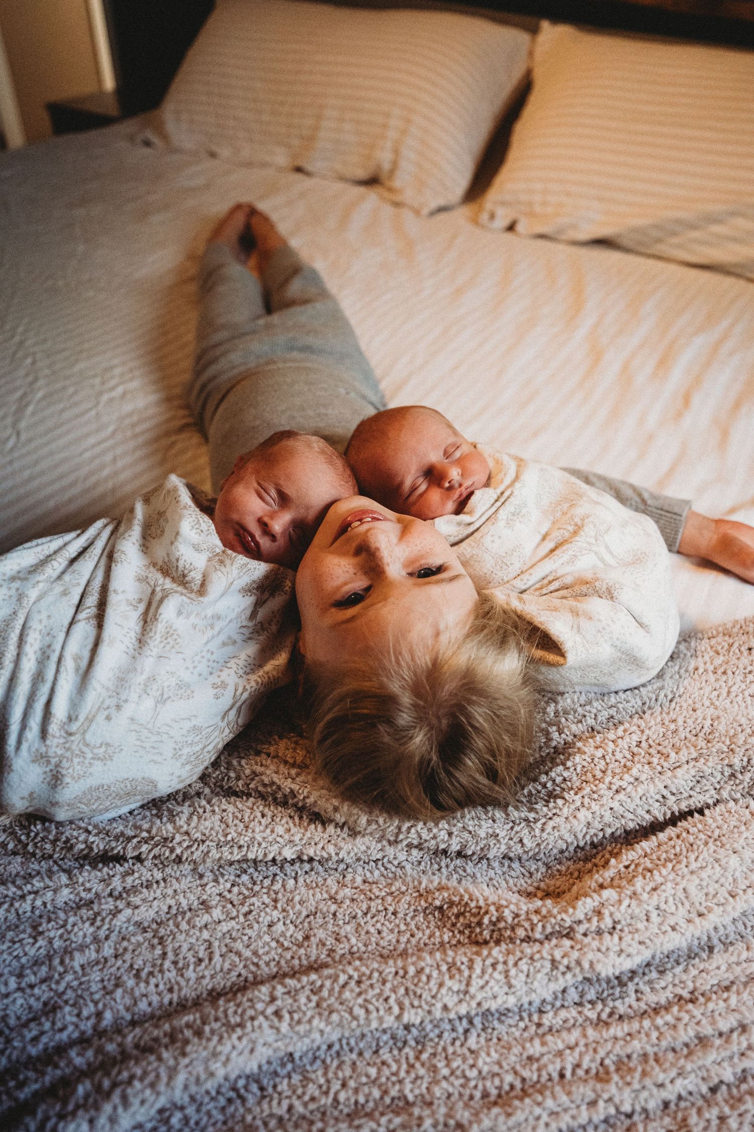 Young child lies on a bed between two sleeping newborns in a warm, intimate family photo.