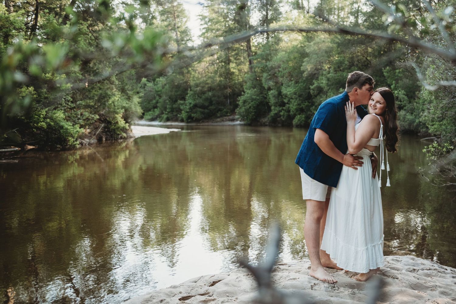 Couple kissing by a serene forest river, man in blue shirt and woman in white dress standing on rocks.